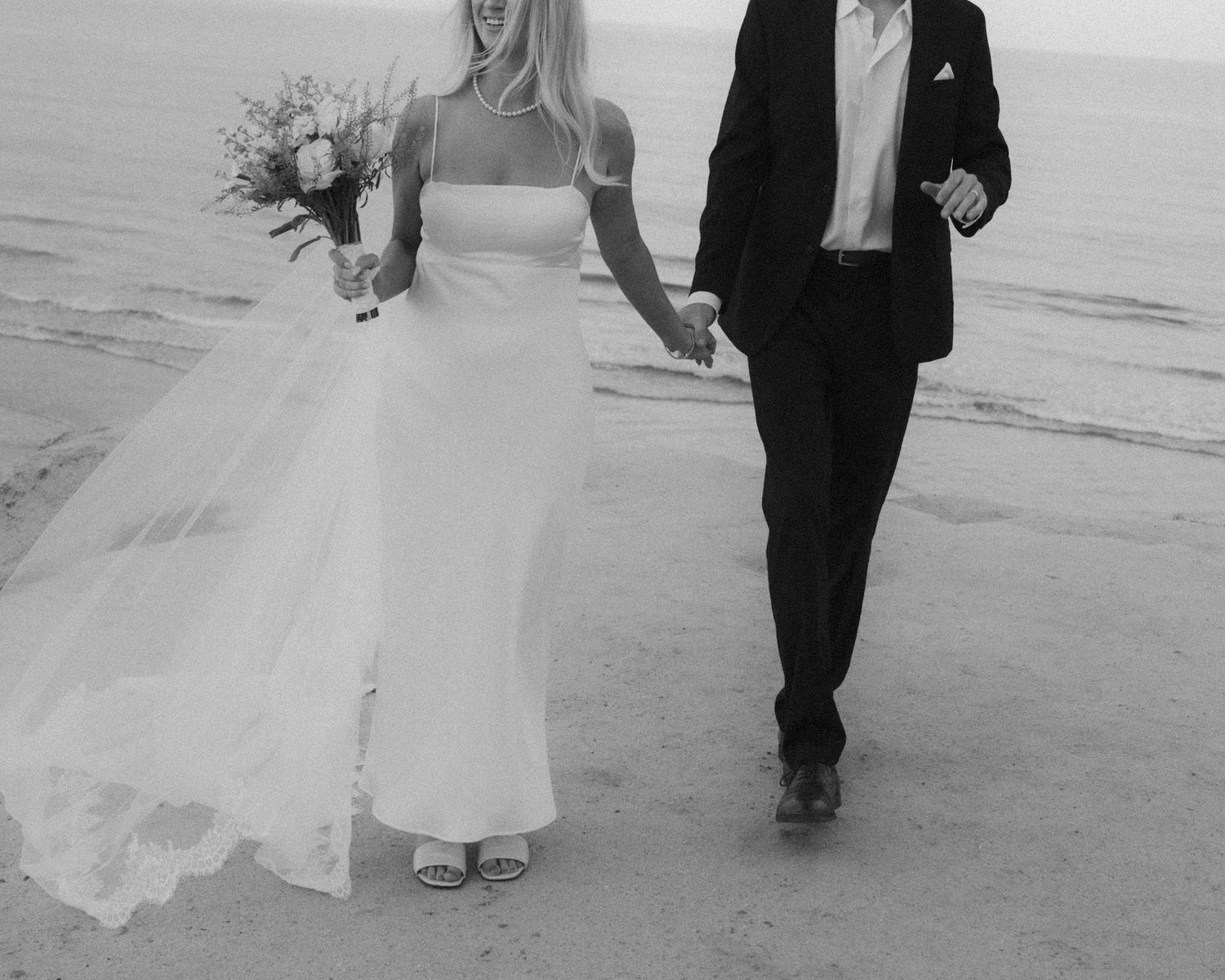 Black and white photo of a bride and groom walking hand in hand on a beach, bride holding a bouquet.