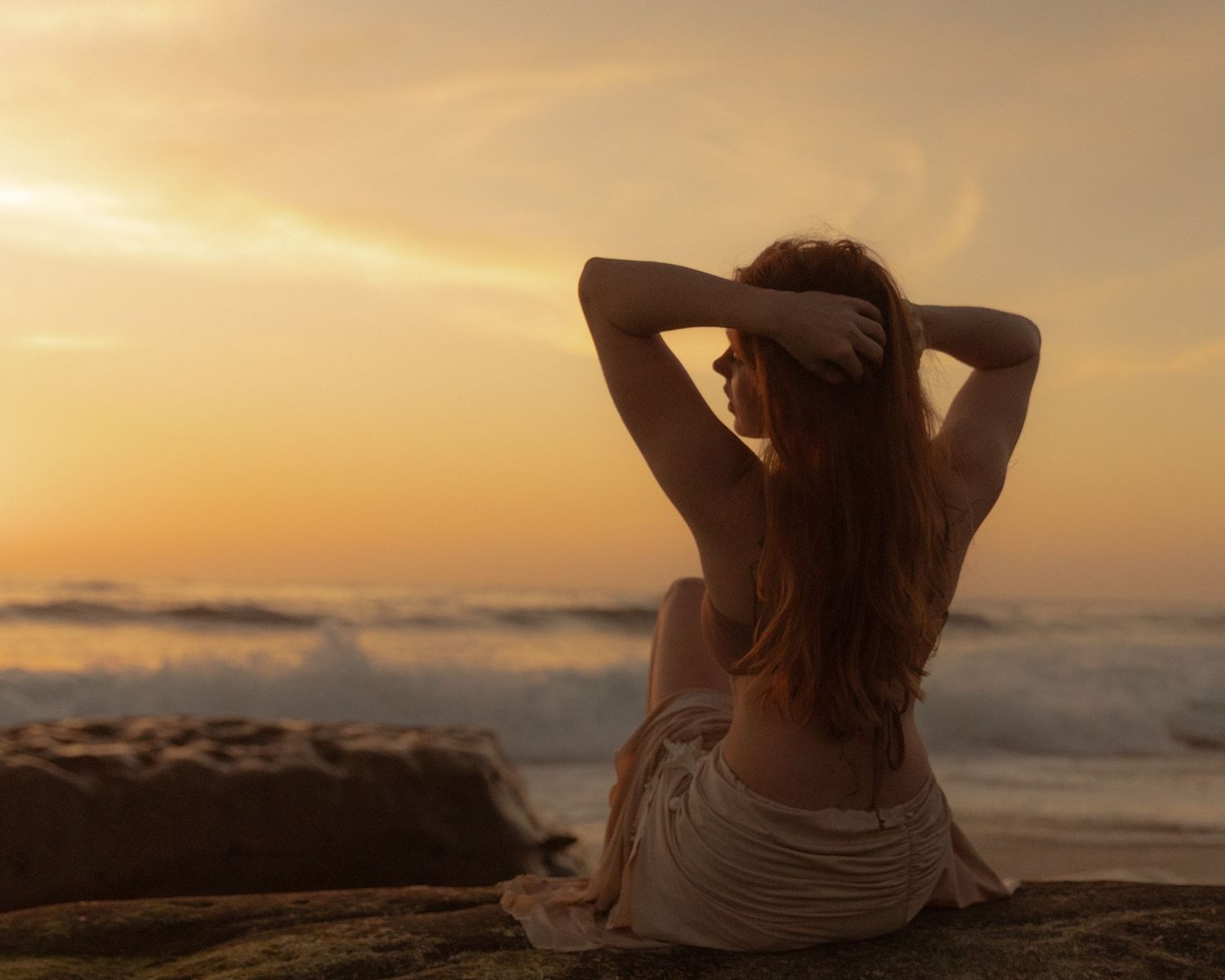 Silhouette of person relaxing on coastal rocks during golden sunset over ocean horizon.