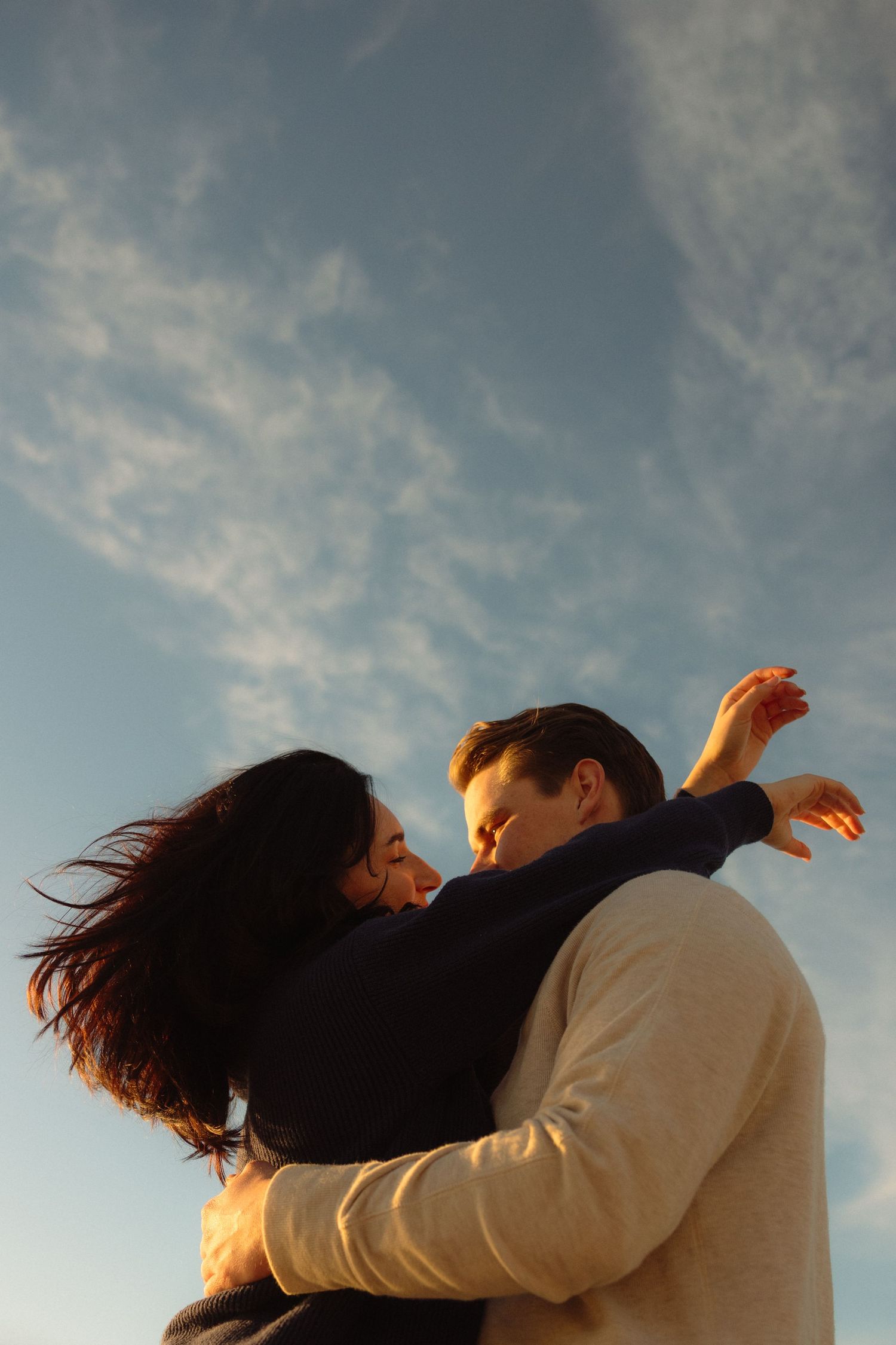 A couple embraces in warm sunset light against a cloudy blue sky.
