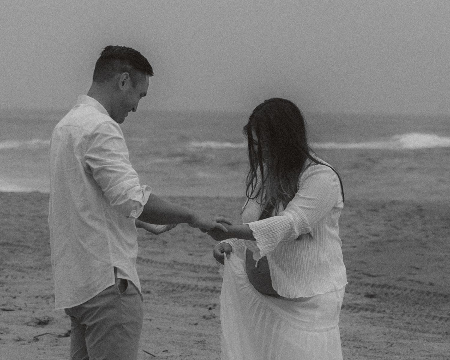 A couple in white clothing embraces on a misty beach as waves crash behind them in a romantic black and white scene.