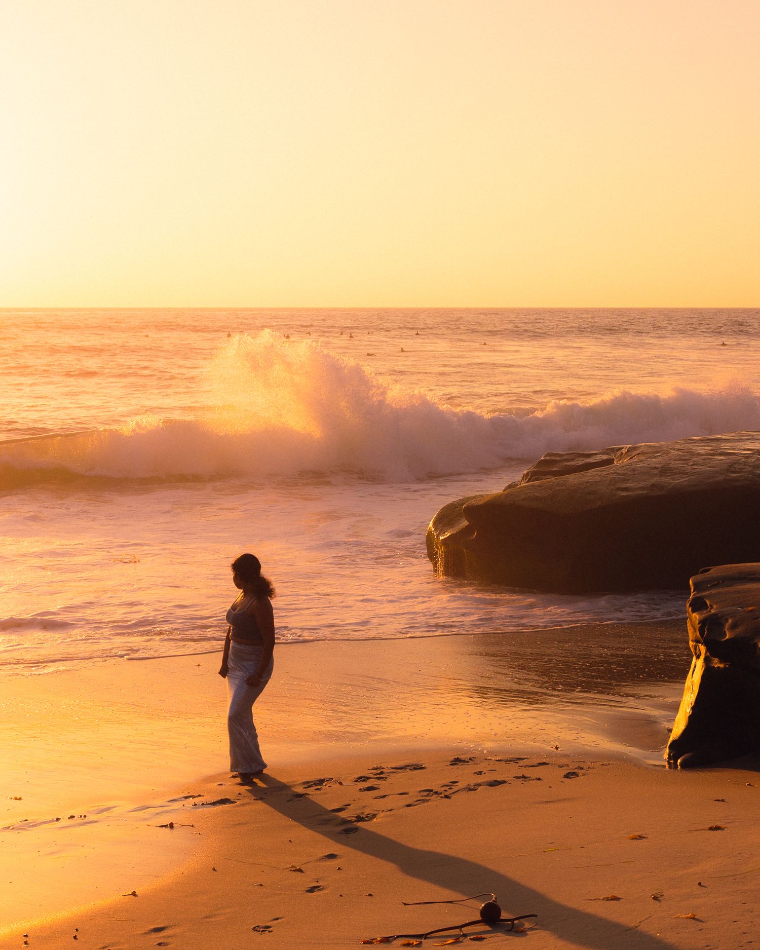 A person walks along a golden beach at sunset while waves crash against rocks creating a dreamy coastal scene.