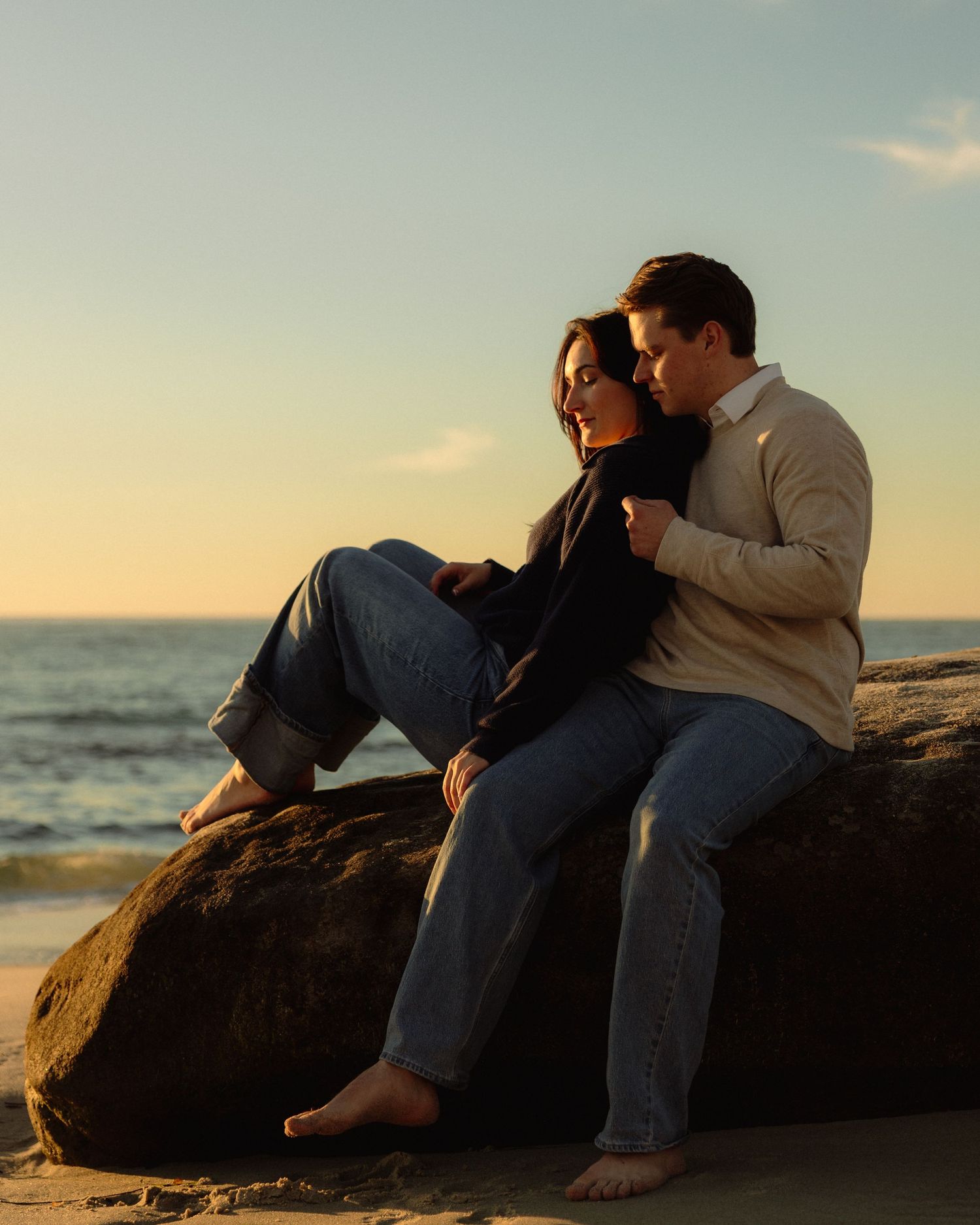 Couple sits together on coastal rocks during sunset with ocean waves crashing in the background.
