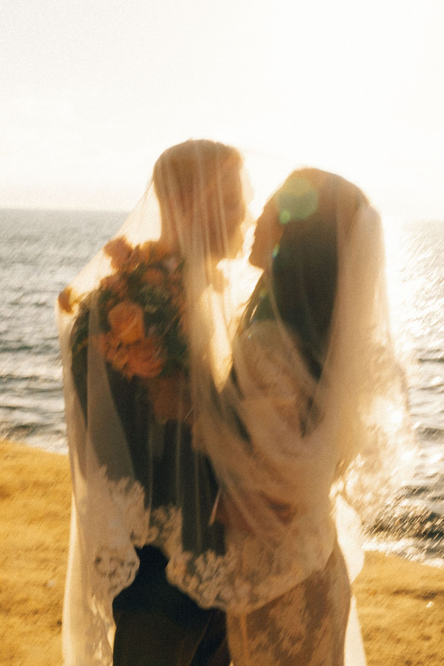 A bride and groom share a romantic kiss under a veil by the ocean at sunset, with a bouquet of orange flowers.