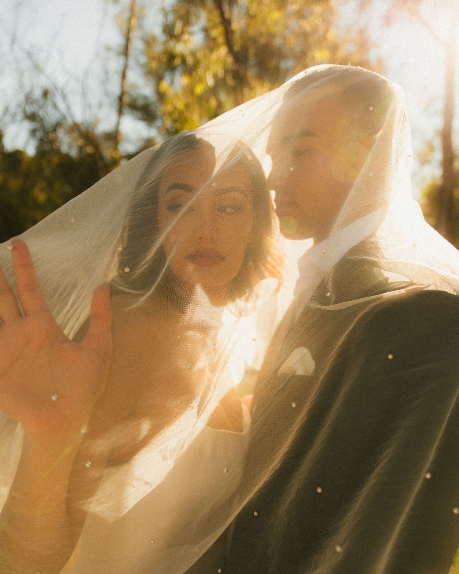 Bride and groom sharing a romantic moment under a sheer veil in golden sunlight outdoors.