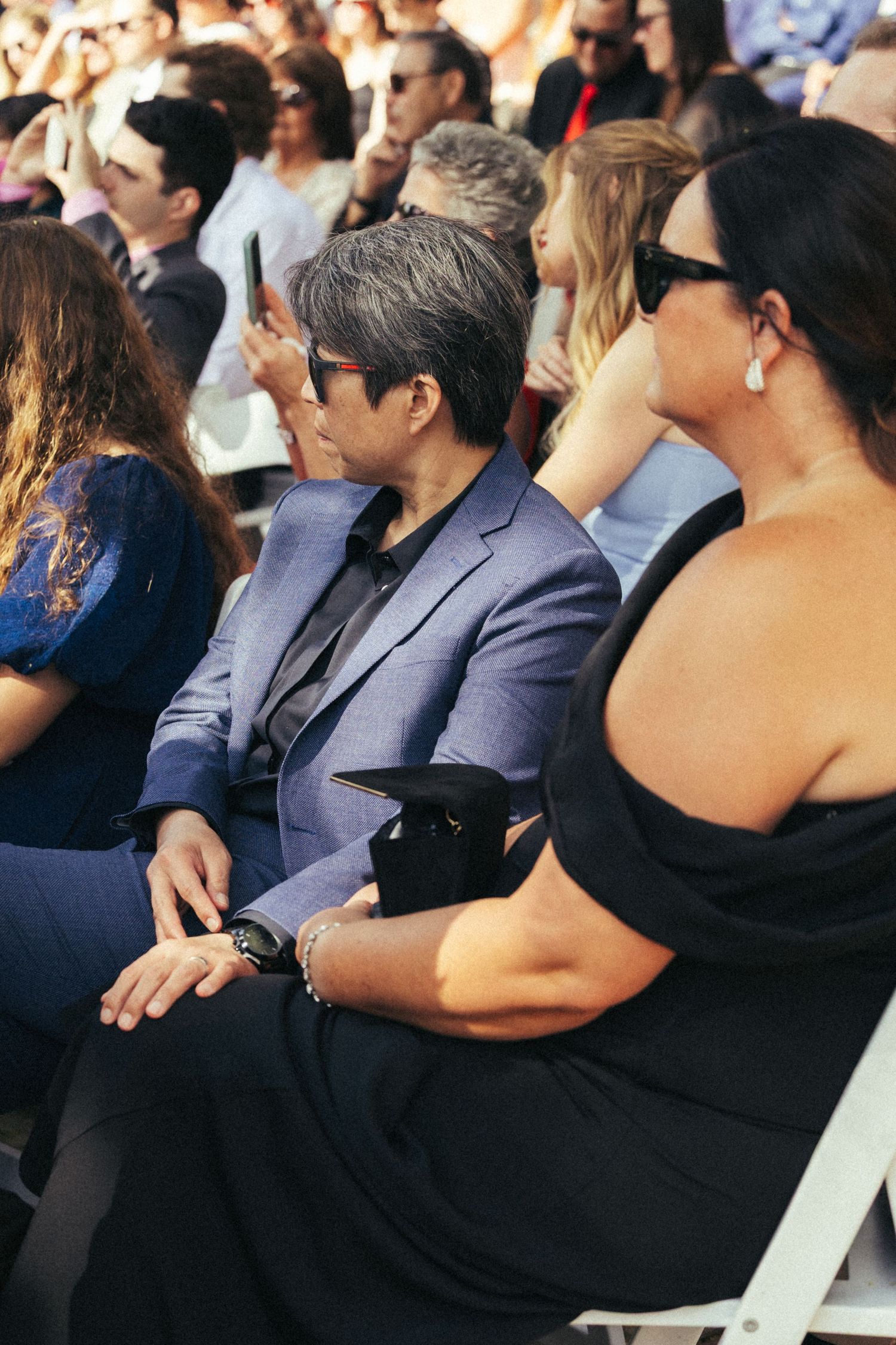 Audience members in formal attire seated at an outdoor event, woman in black dress holding clutch purse.