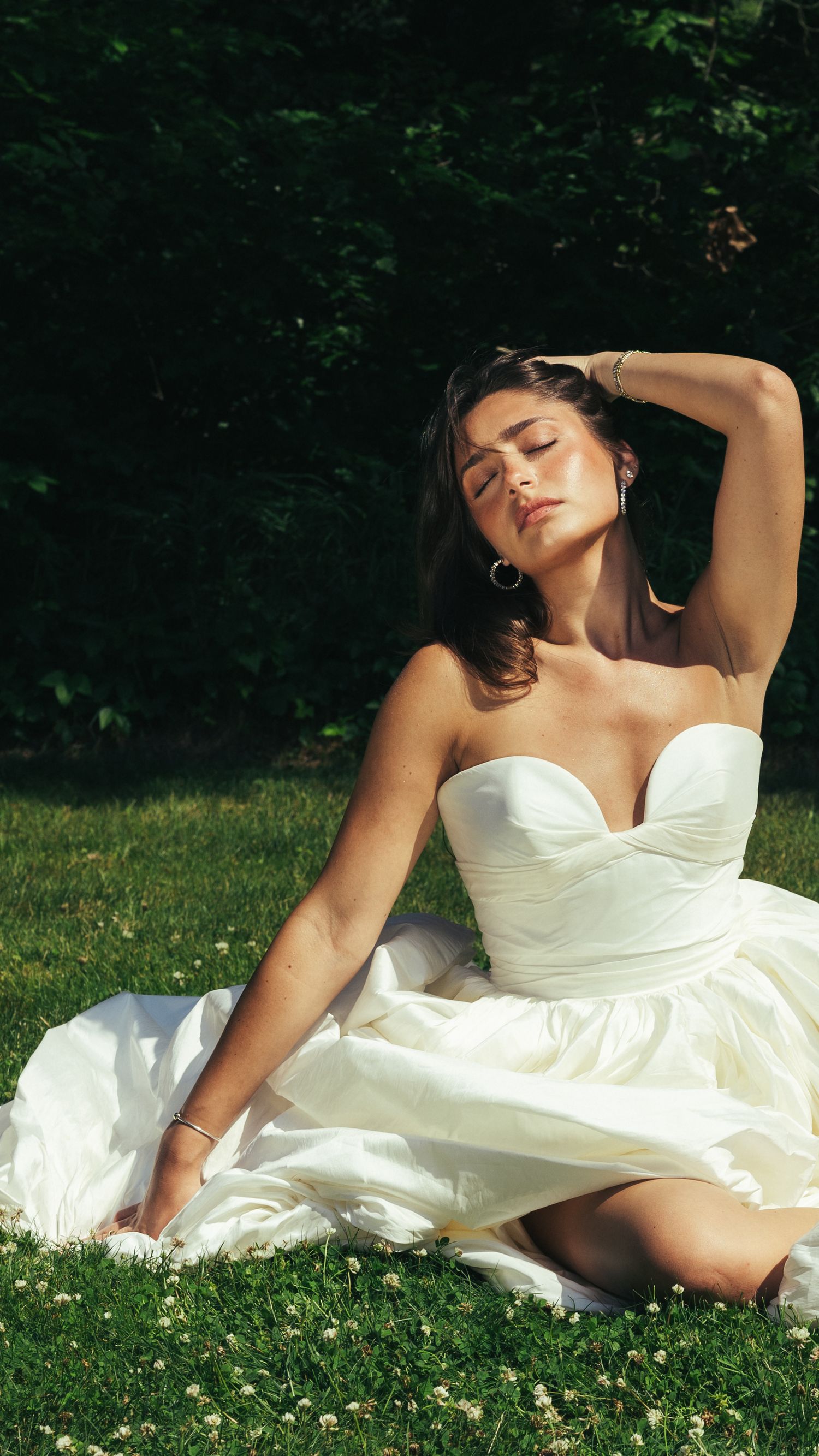 Woman in strapless white ball gown sitting on grass with eyes closed in sunlight.