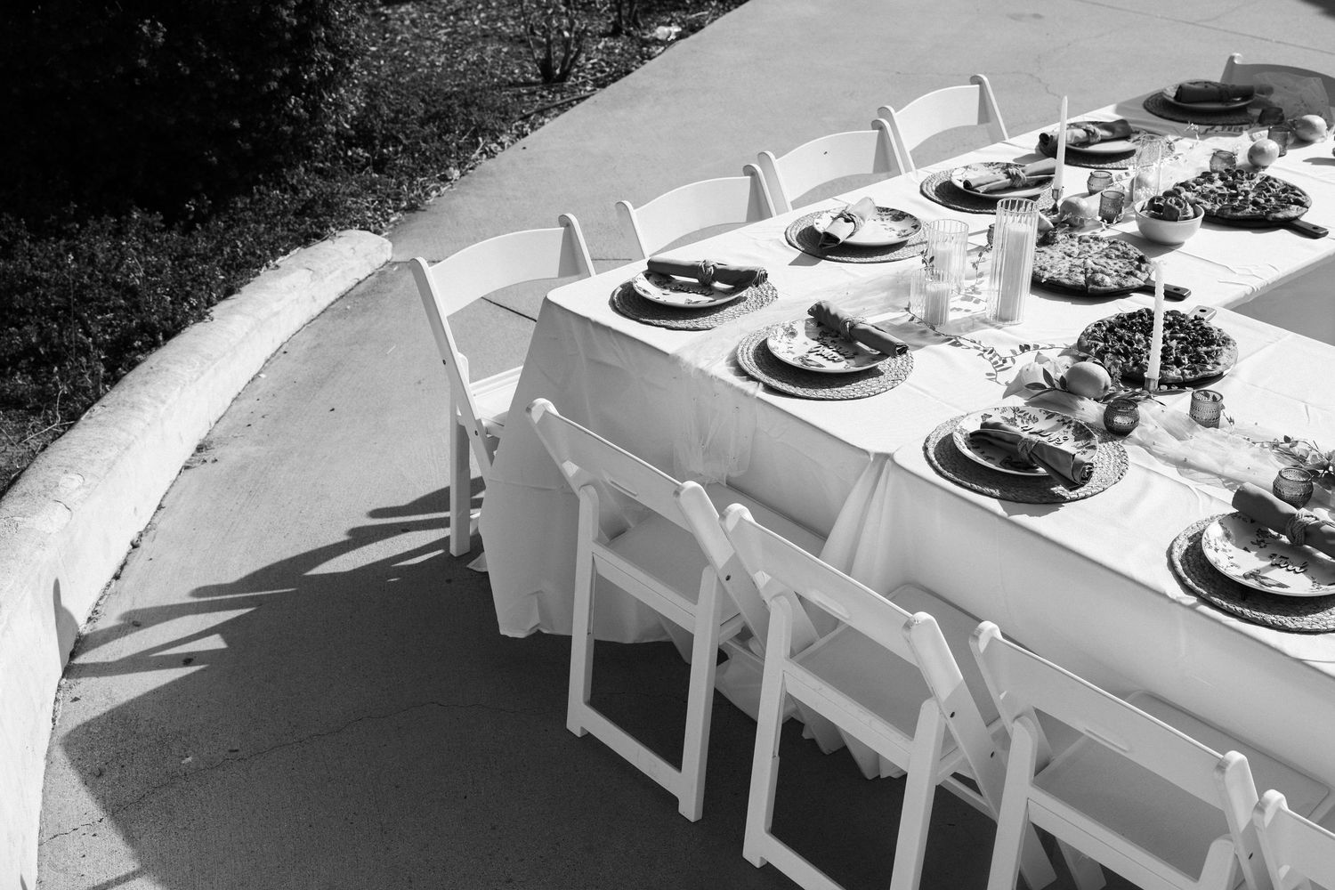 Black and white photo of an elegant outdoor dining table set with dark plates and floral centerpieces, surrounded by white folding chairs.