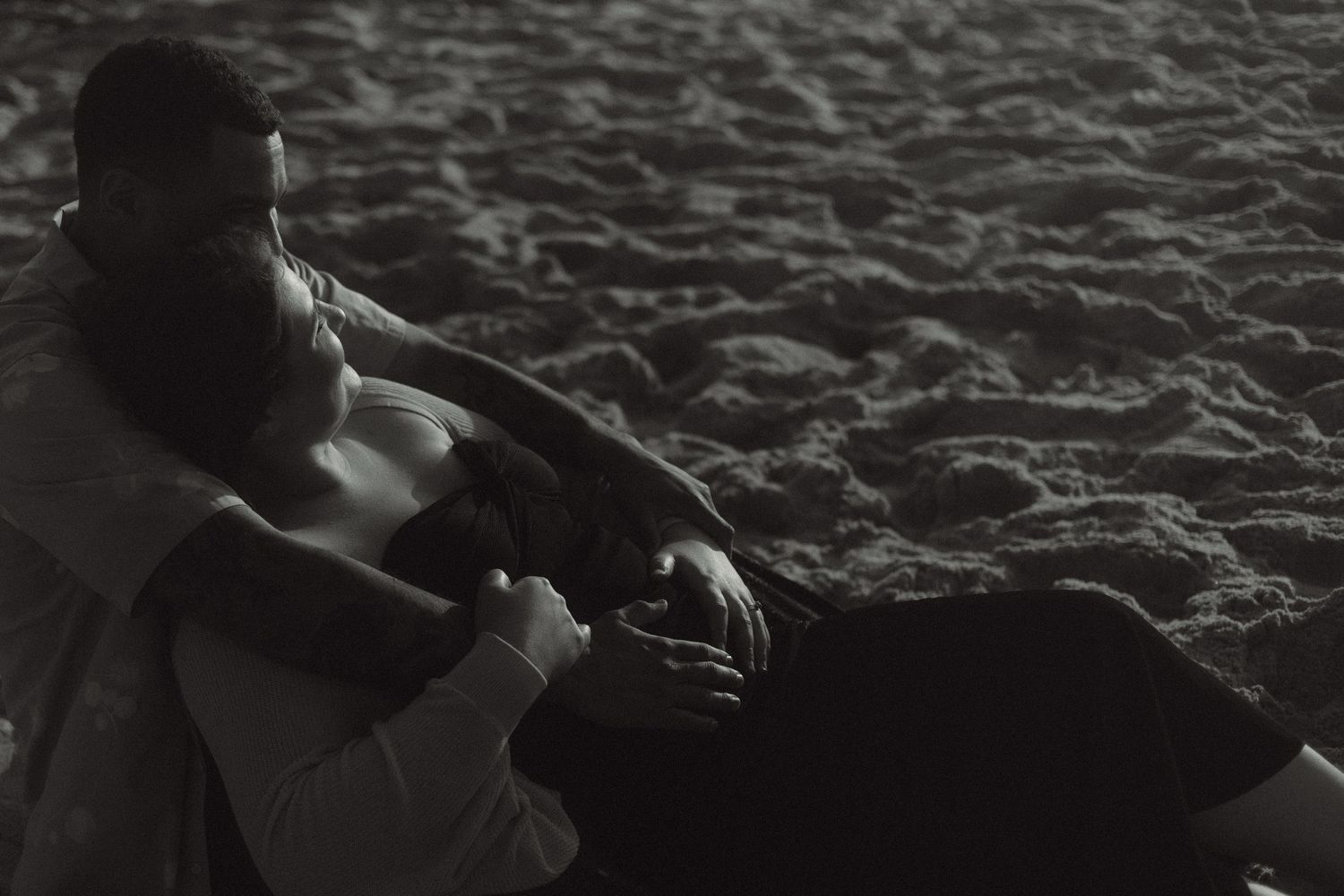 A couple sits together on sandy beach at night in an intimate black and white moment.