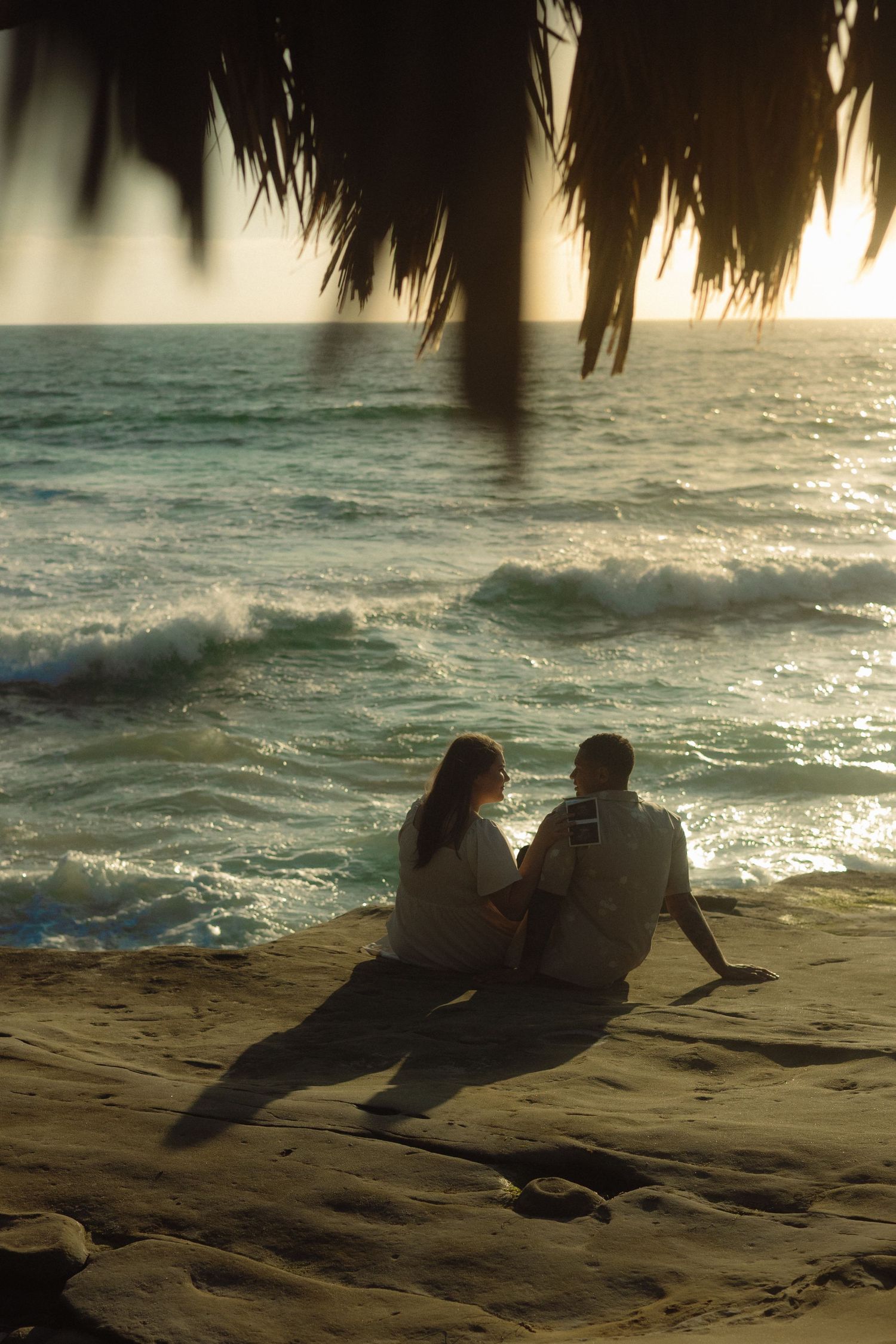 A couple sits silently on a beach at sunset, framed by palm fronds overhead as waves roll onto the shore.