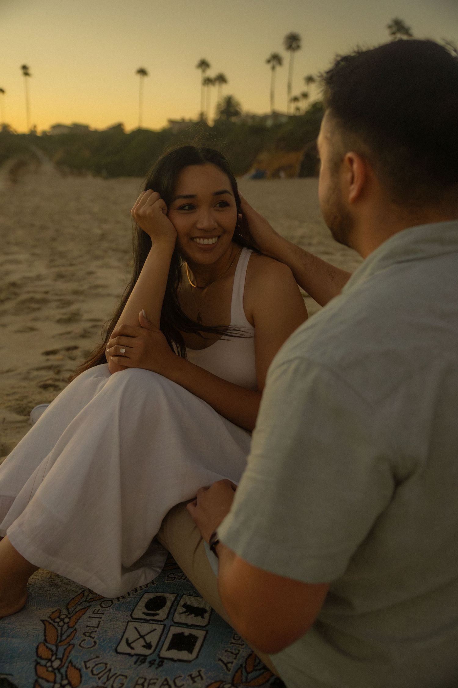 A couple shares a tender moment together on a beach at sunset with palm trees in the background.
