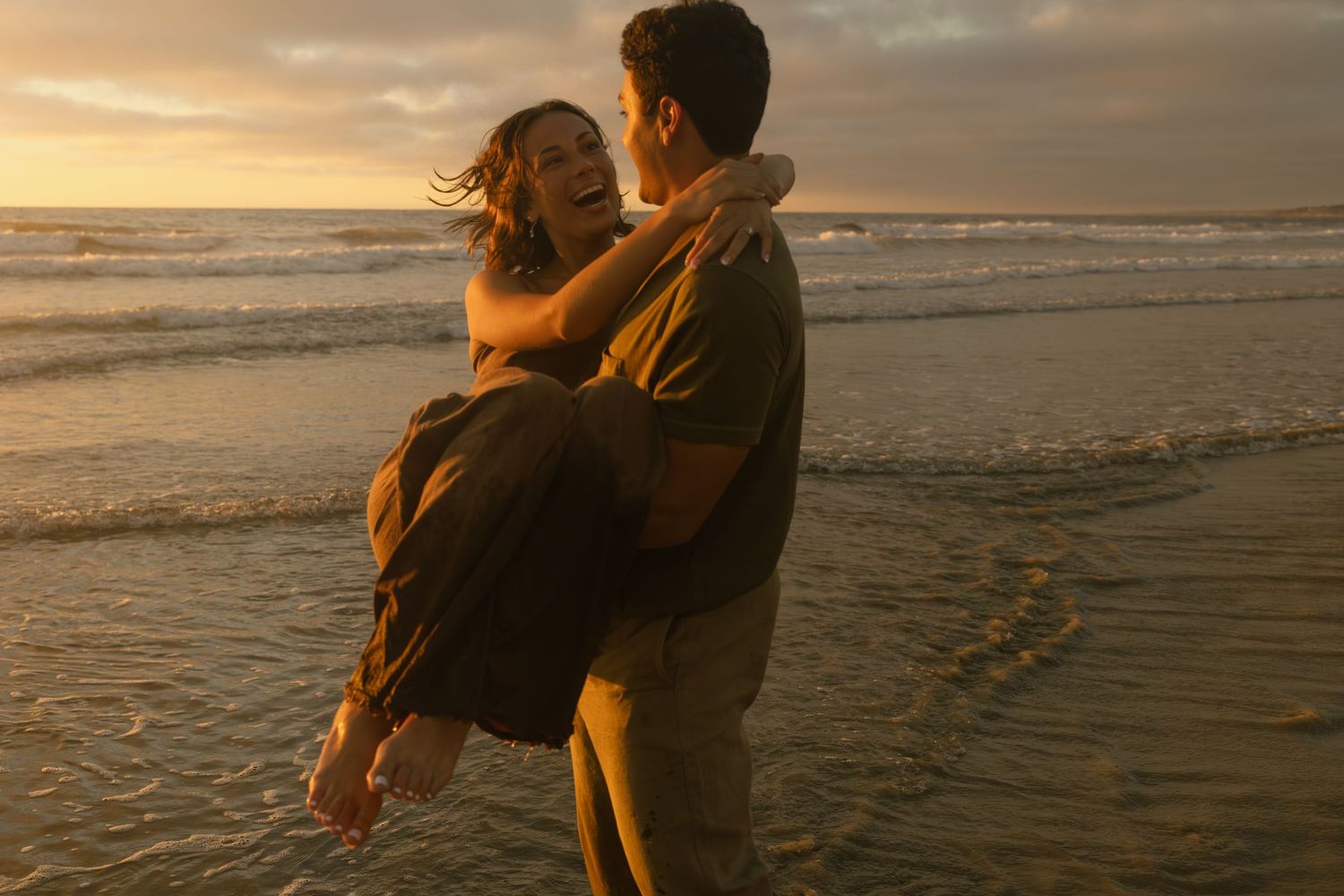 Couple embraces on sandy beach at sunset as waves roll onto shore in romantic seaside moment.