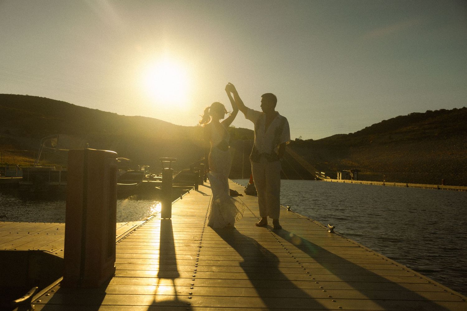 Silhouetted figures walk along a waterfront pier at sunset with golden sunlight reflecting on the calm water.