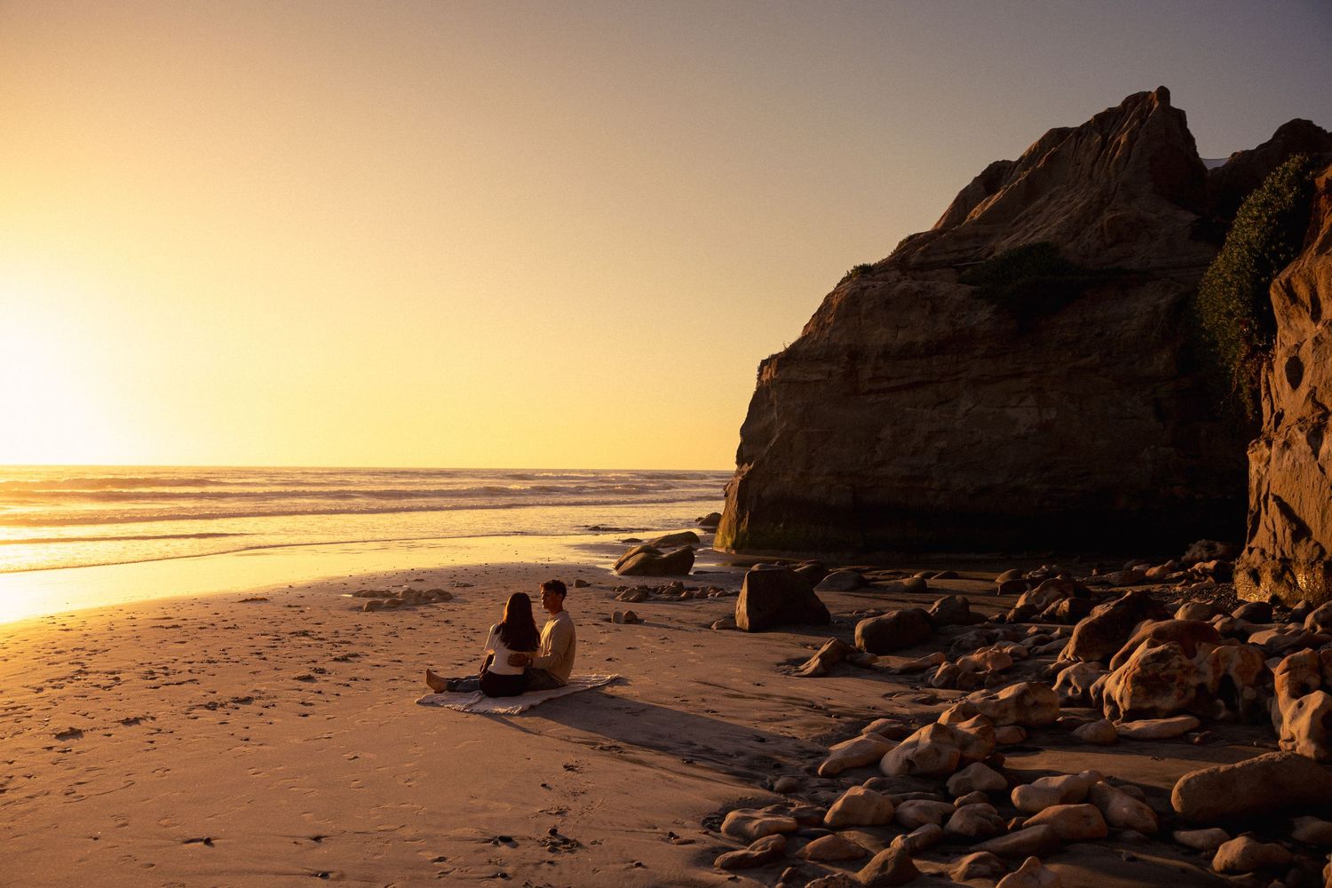 Golden sunset illuminates a rocky beach with dramatic cliffs and scattered boulders along the Pacific coast.