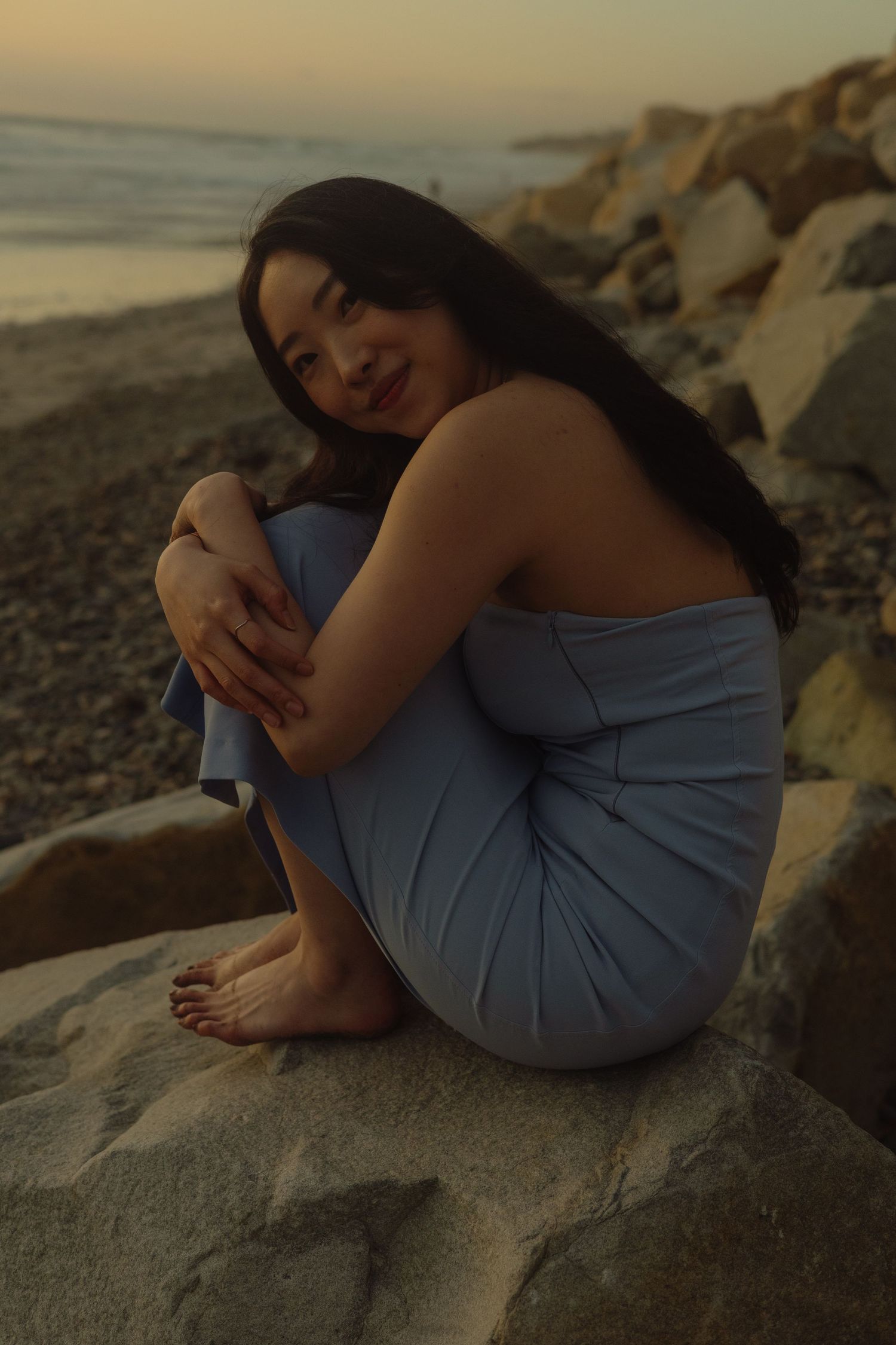 Woman in blue dress reclining on coastal rocks during golden hour.