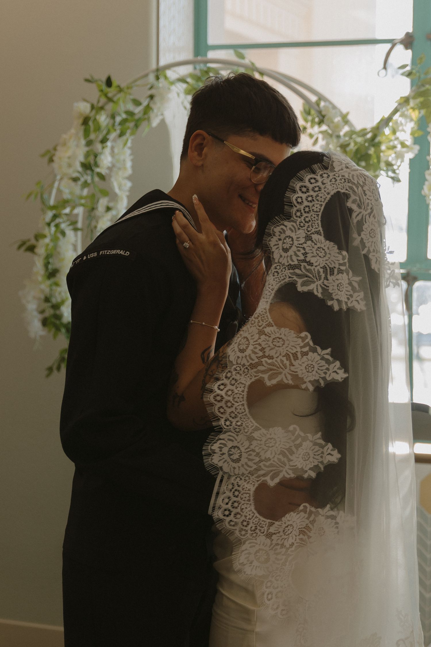 A bride in lace veil embraces a sailor in dress uniform during an indoor wedding ceremony.