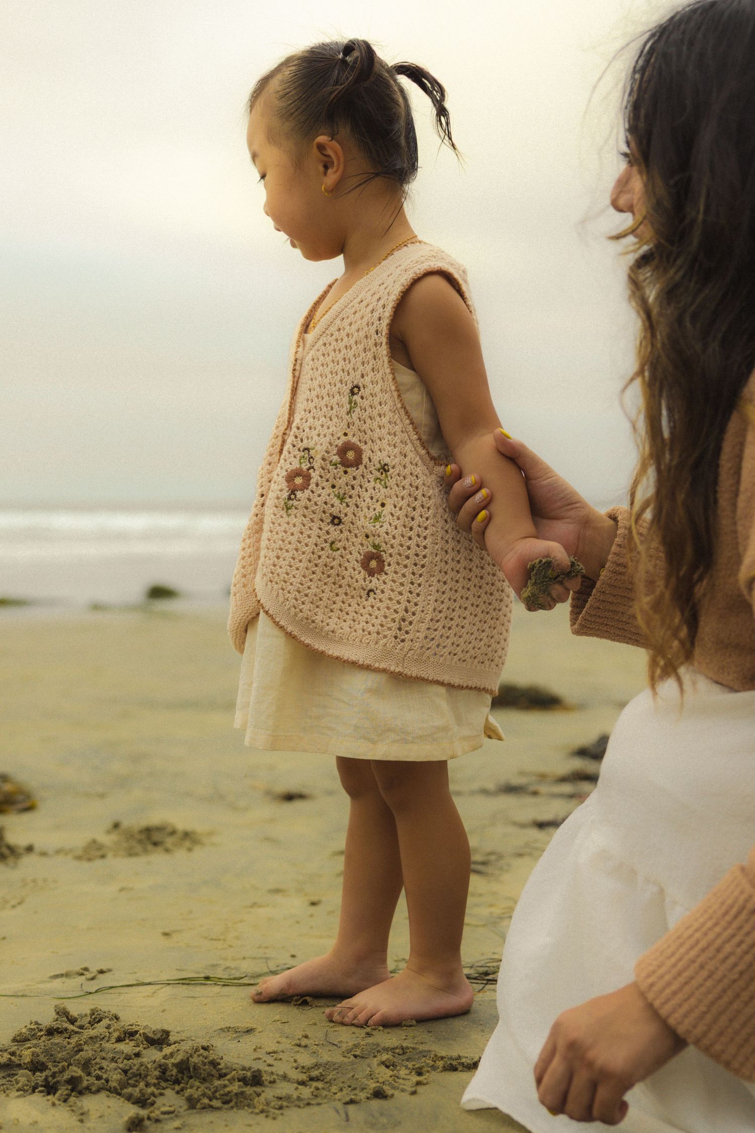 A child in a crocheted beige dress walks along a misty beach with an adult figure visible in the background.