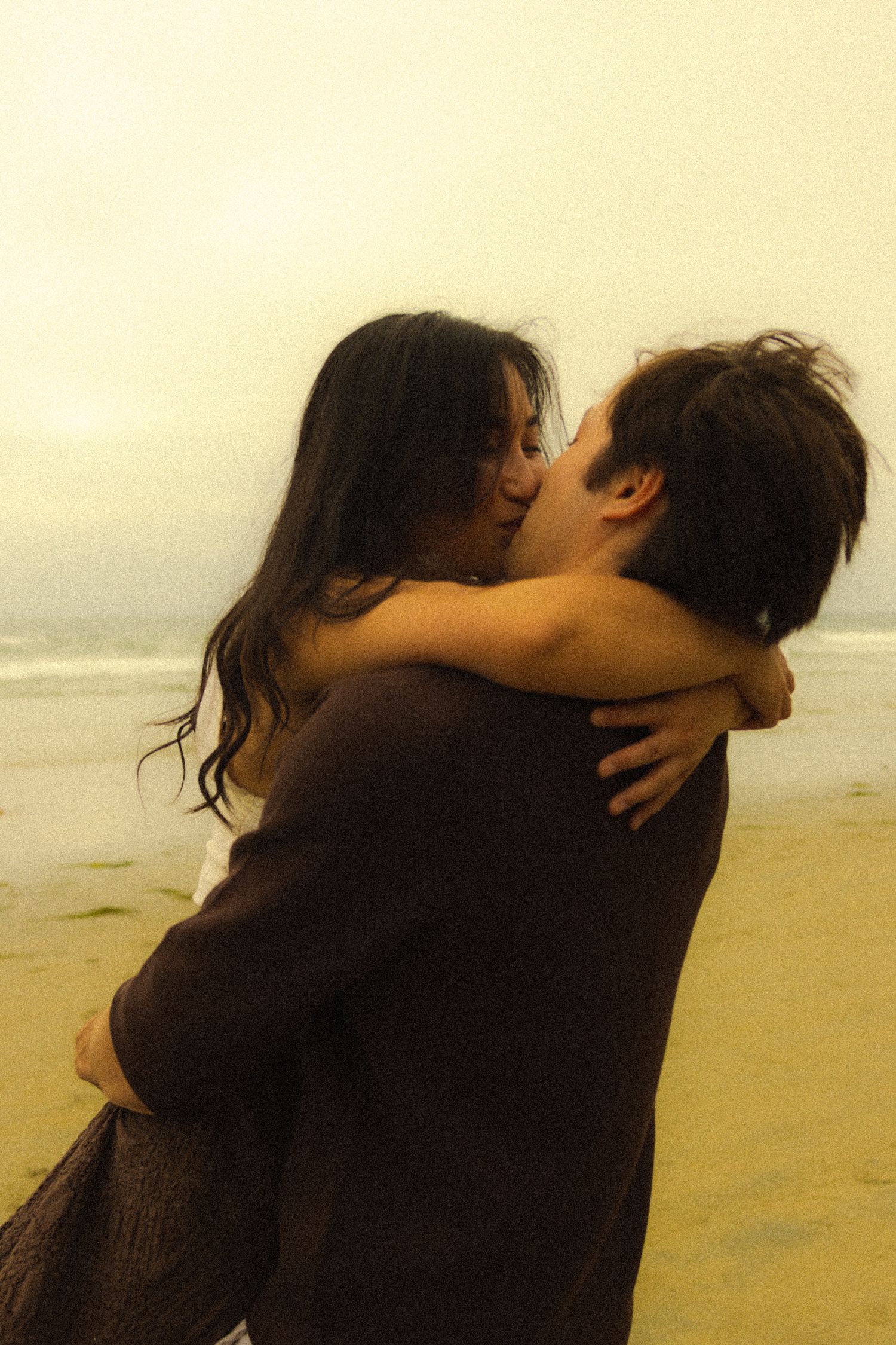 A romantic couple embraces and kisses on a misty beach at sunset with the ocean in the background.