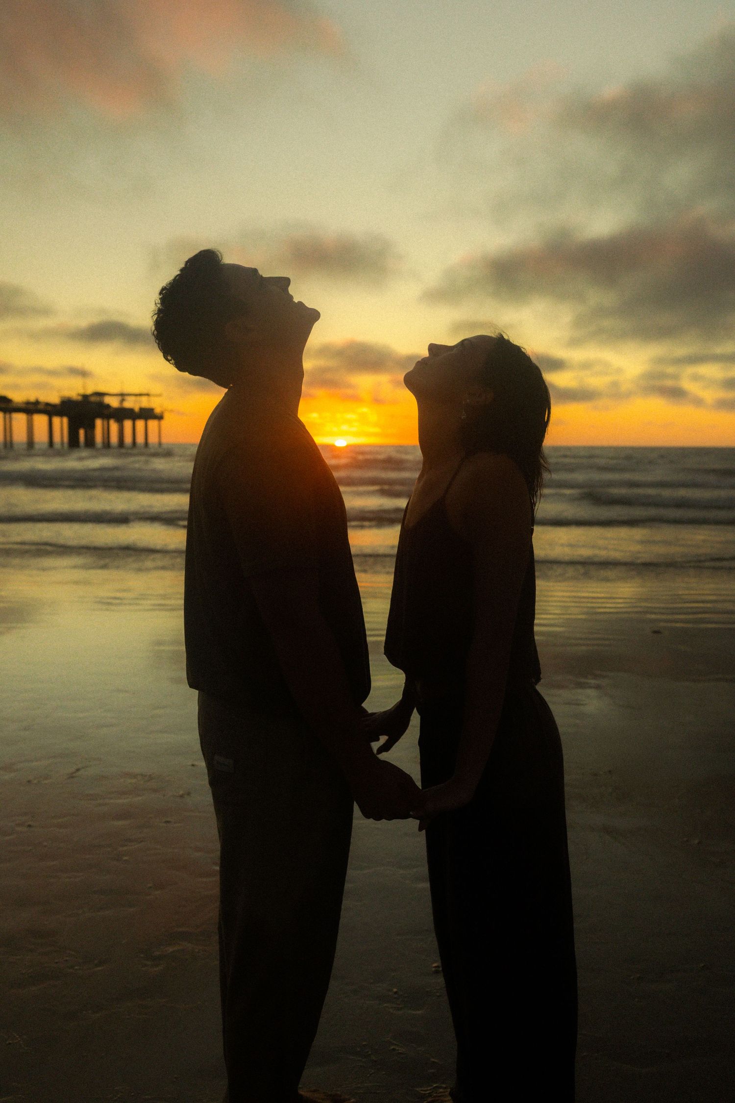 Silhouettes of two figures standing on a beach at sunset with a pier in the distance.