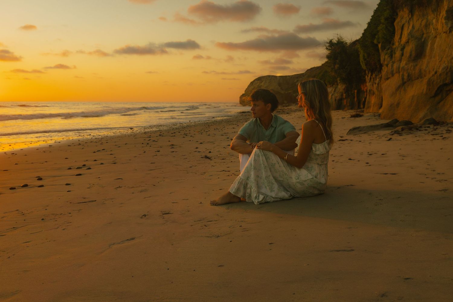 A figure in a flowing dress sits on a sandy beach at sunset beneath towering cliffs along the shoreline.