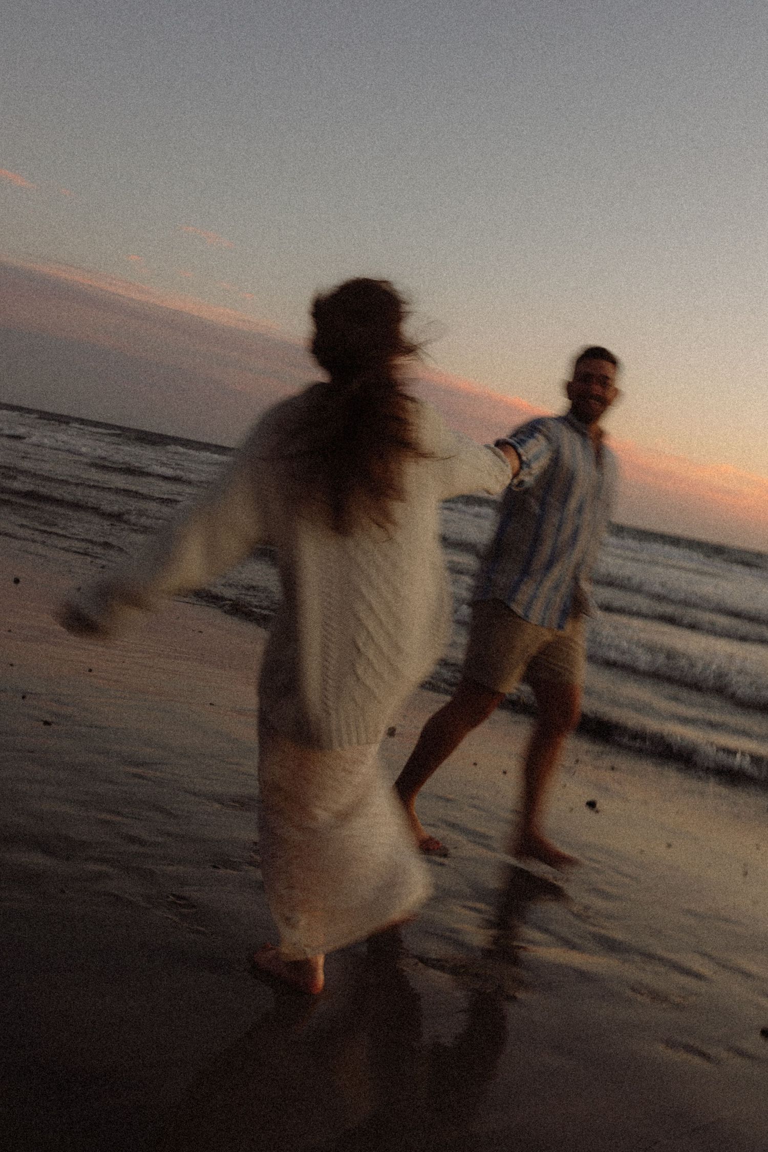 Couple dancing barefoot on a beach at sunset, woman in white knit dress, man in blue shirt and shorts.
