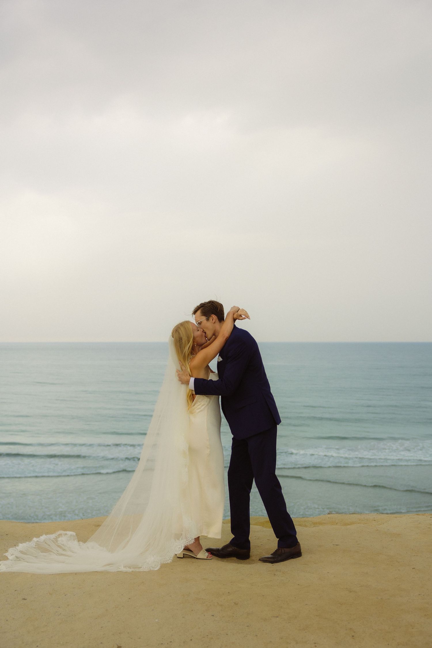 Bride and groom share a romantic kiss on a sandy beach, with turquoise ocean waves and overcast sky behind them.
