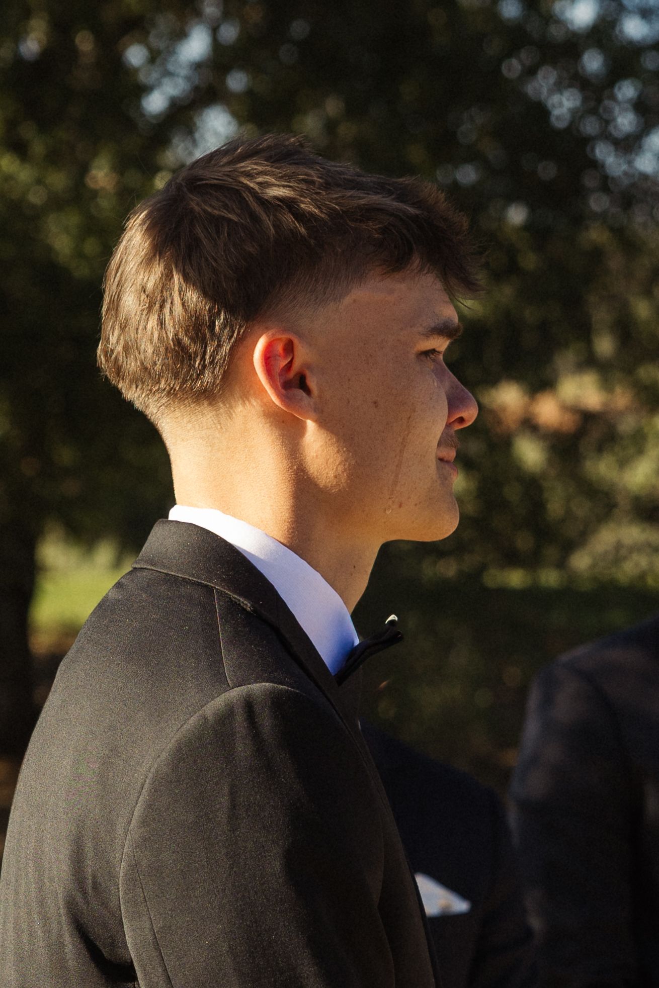 Young man in dark suit jacket with short hair, photographed in profile outdoors with trees in background.
