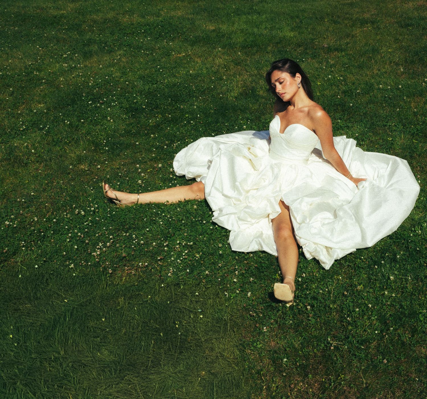 Bride in a white strapless ball gown sitting barefoot on lush green grass, with her dress spread around her.