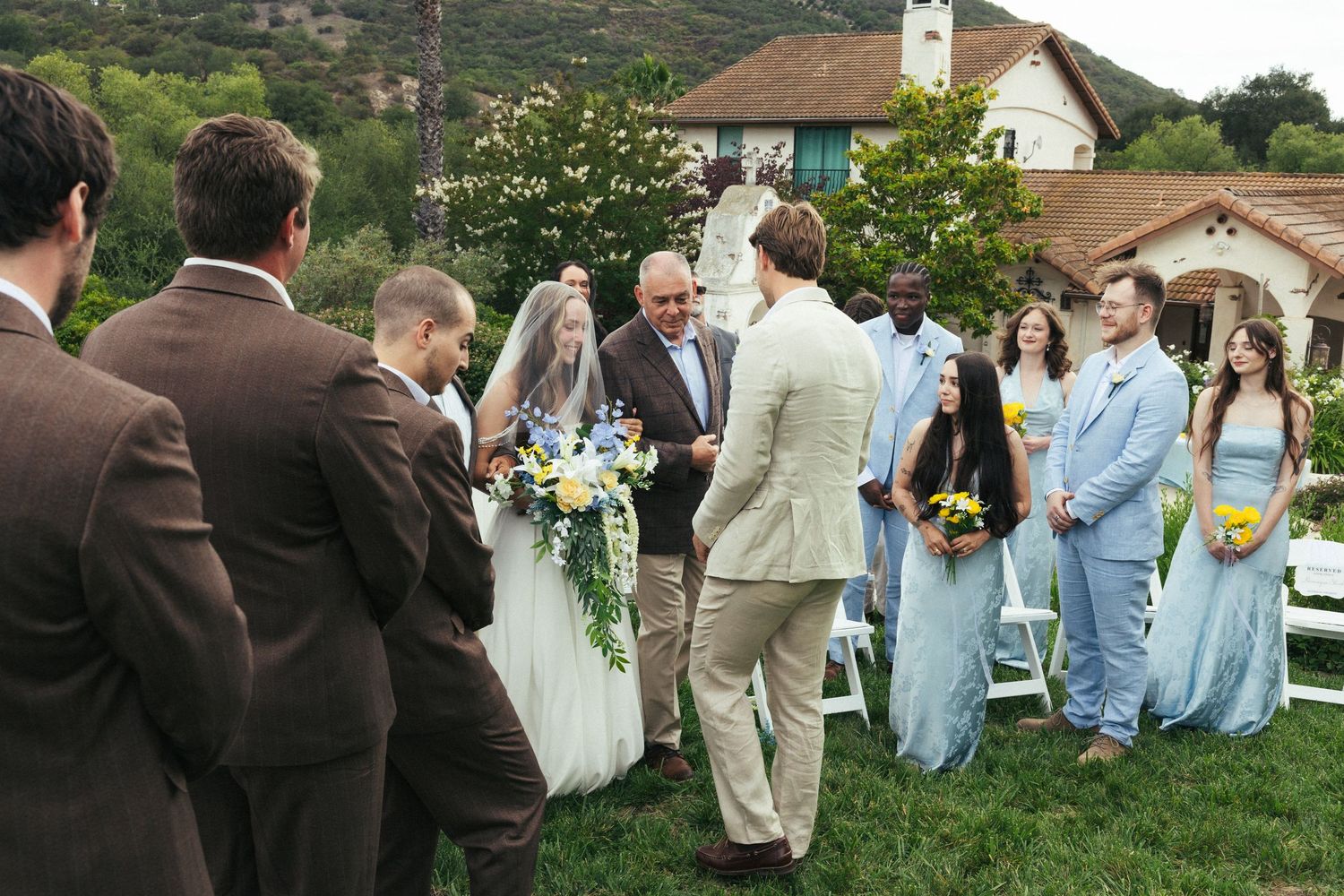 Outdoor wedding ceremony on green lawn with bride, groom, officiant, groomsmen in brown suits, and bridesmaids in blue dresses.