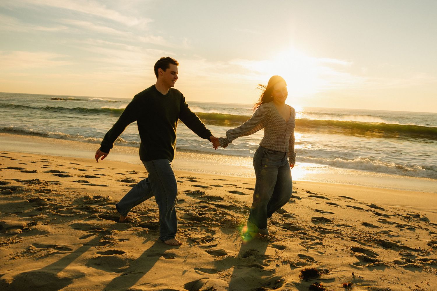Silhouetted figures playfully run and hold hands along a beach at golden sunset with ocean waves in background.