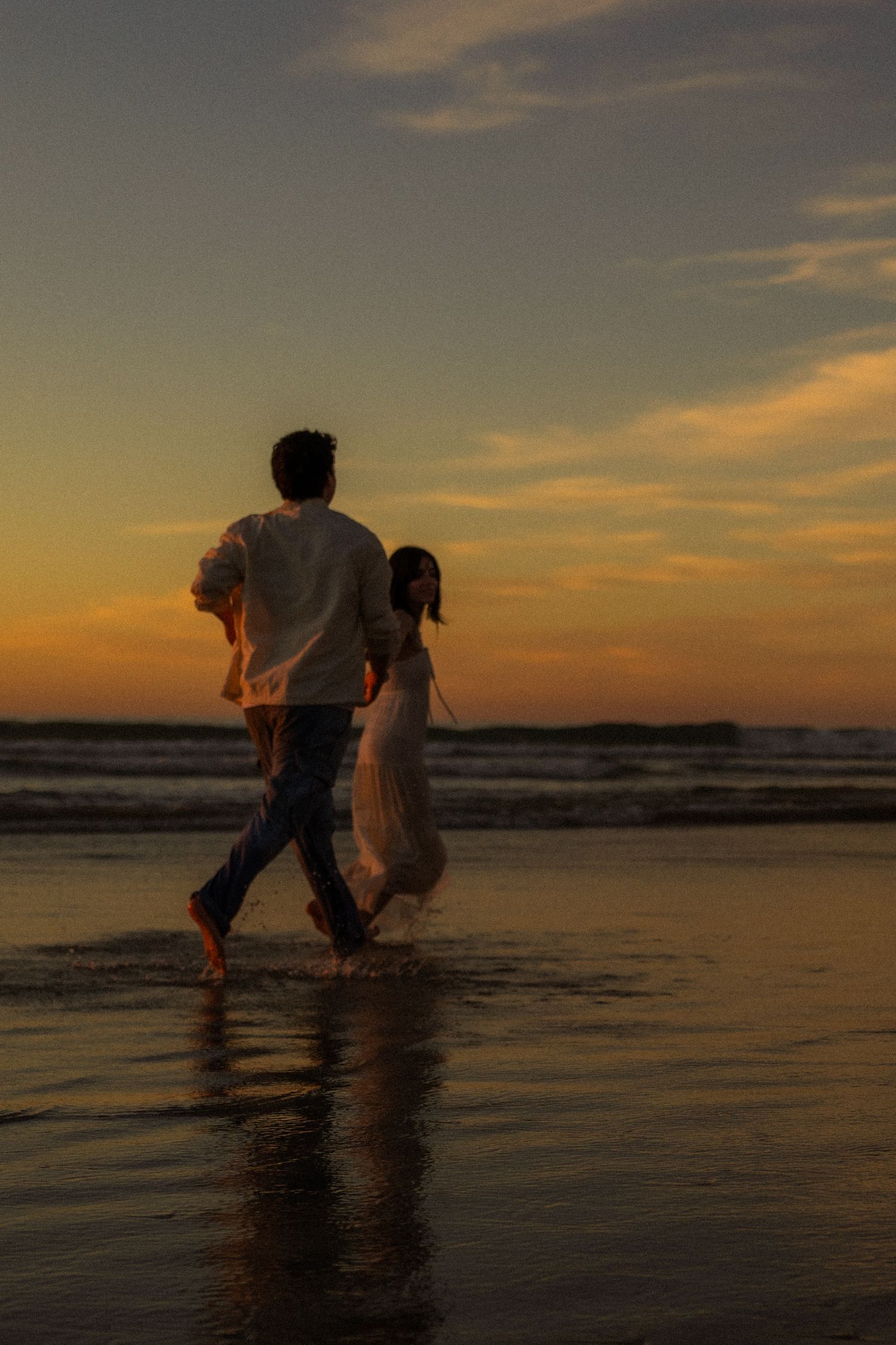 Two silhouettes walk together along a beach at sunset with golden sky reflecting on wet sand.