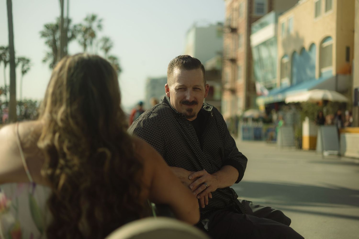 Two people sit having an animated conversation at an outdoor cafe table on a sunny urban street.