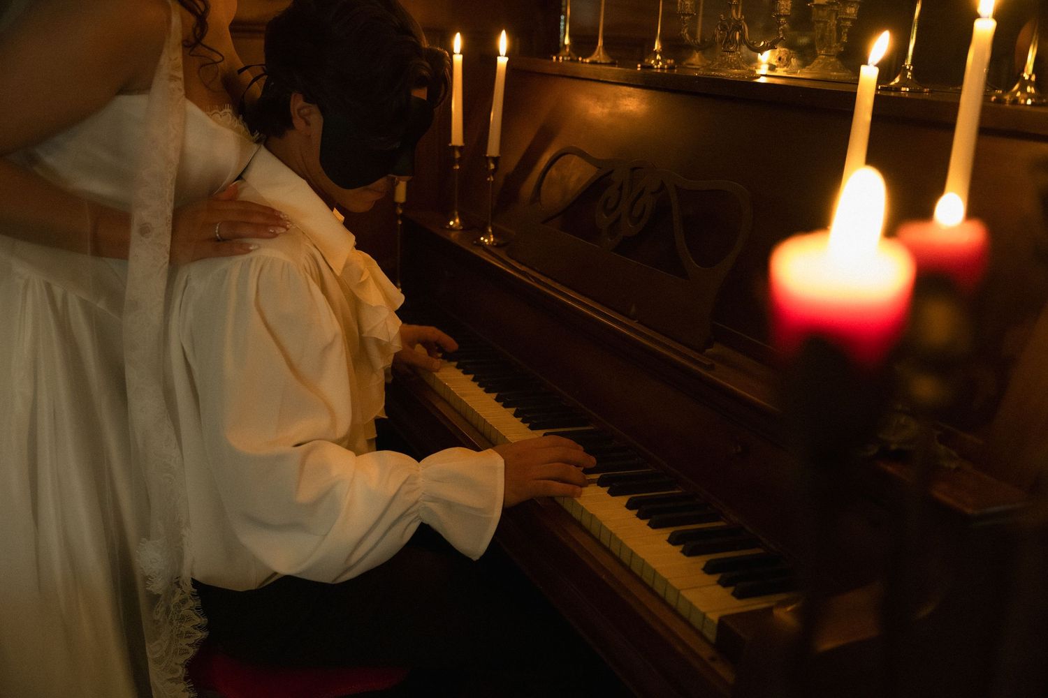 A pianist in white clothing plays by candlelight on a grand piano in a dimly lit room.
