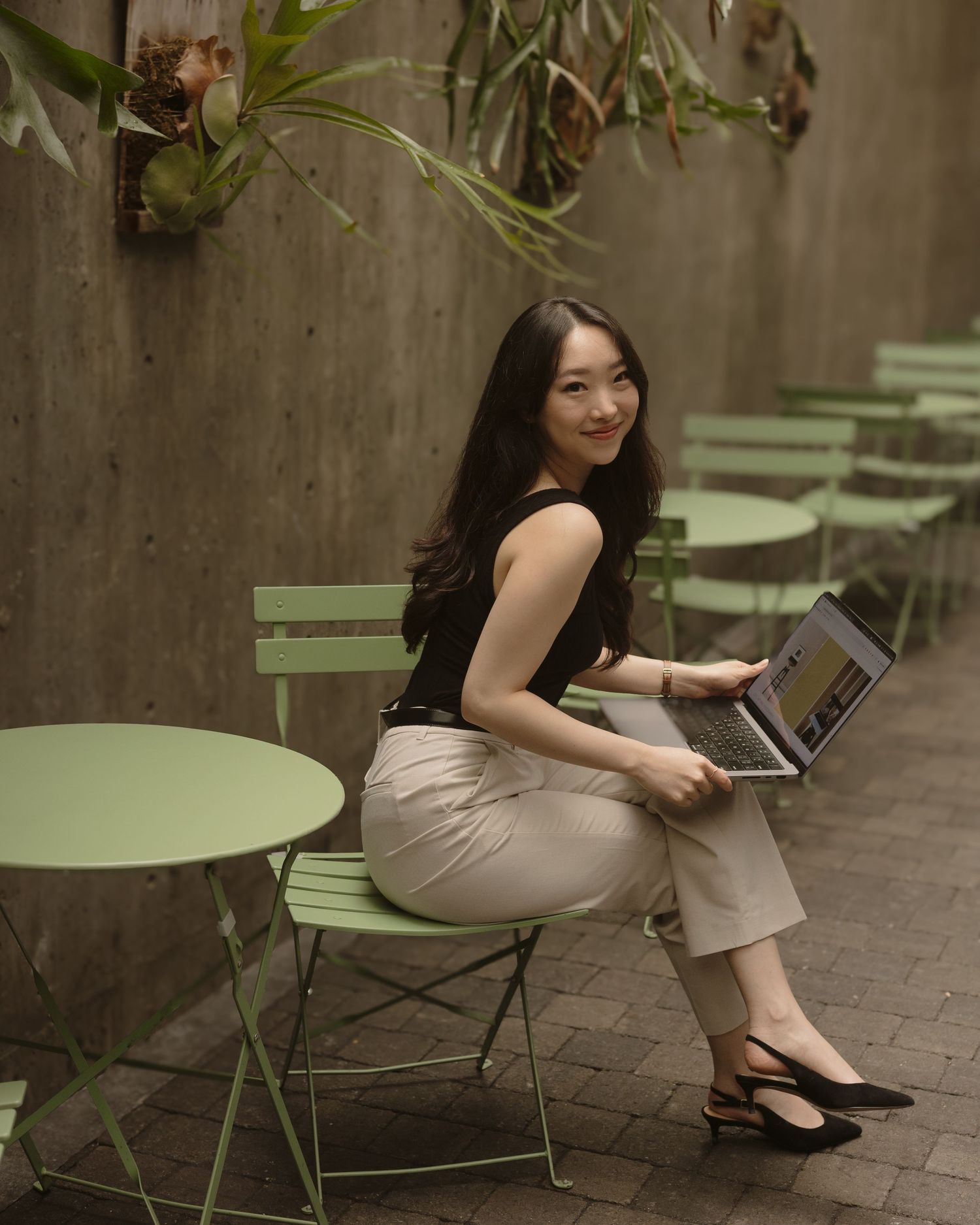 A woman in business casual attire sits at a green outdoor cafe table while working on a laptop computer.