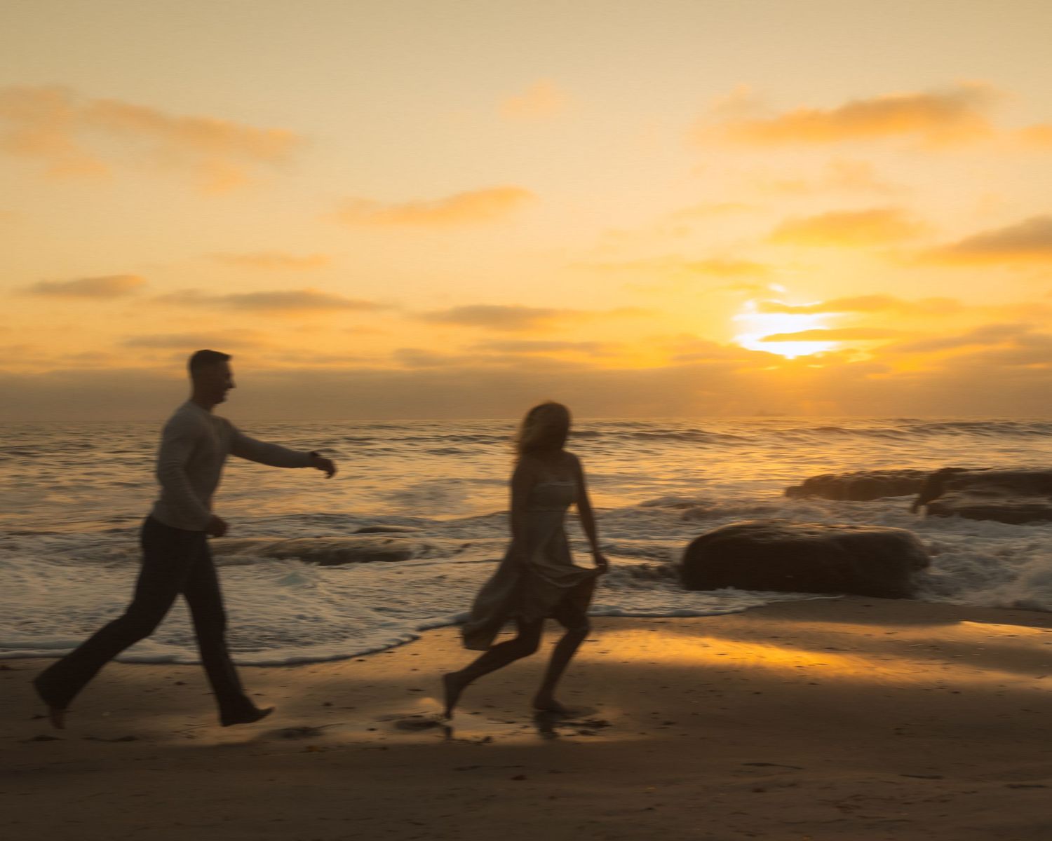 Silhouetted figures dance playfully along a beach at sunset with golden orange sky and rolling waves in background.