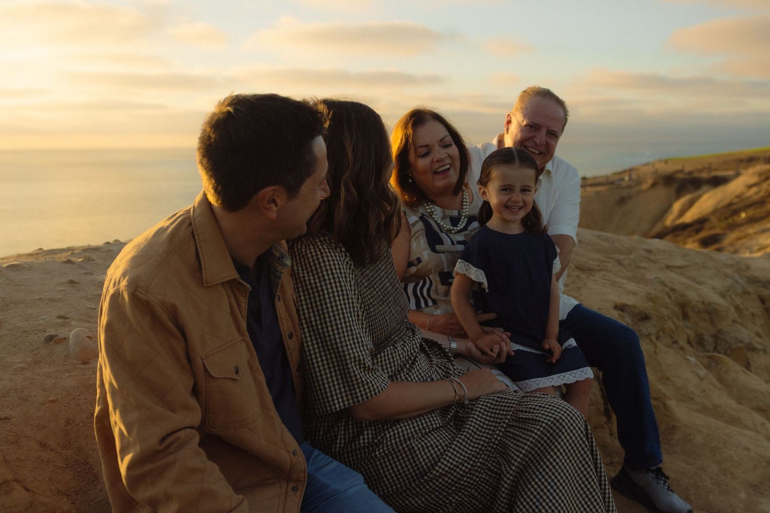 A group of people gather on a sandy beach dune during golden hour as the sun sets over the horizon.