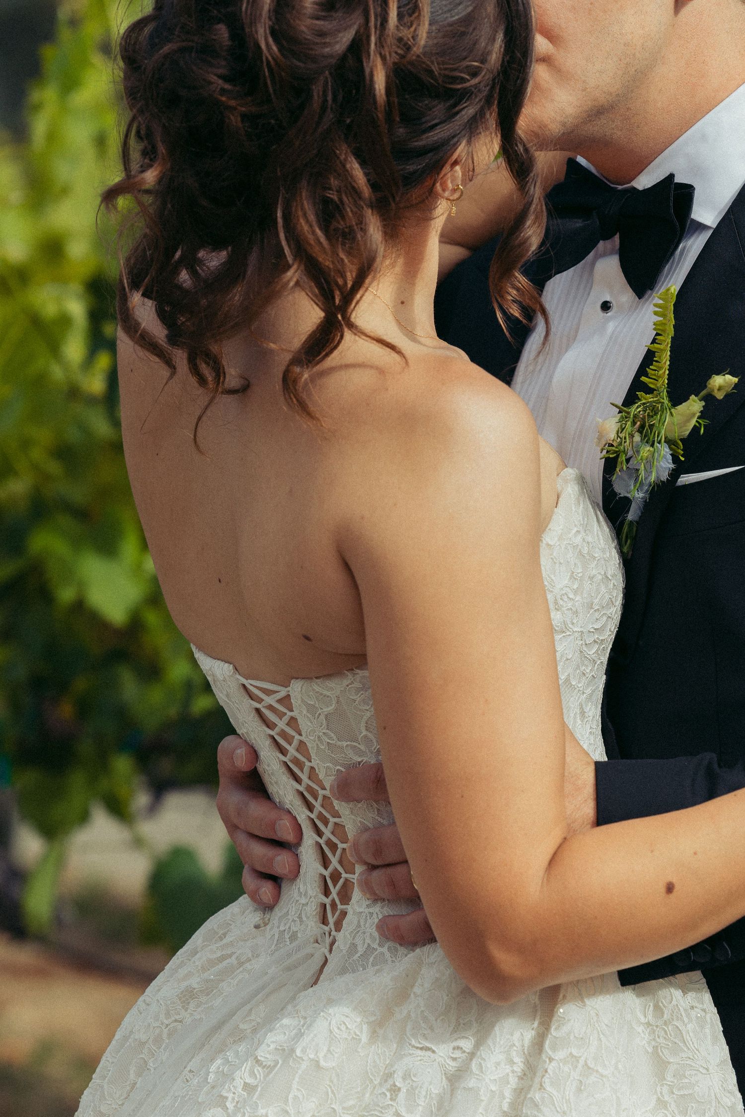 Bride in lace strapless gown embraced by groom in black tuxedo with boutonniere, sharing a kiss outdoors.