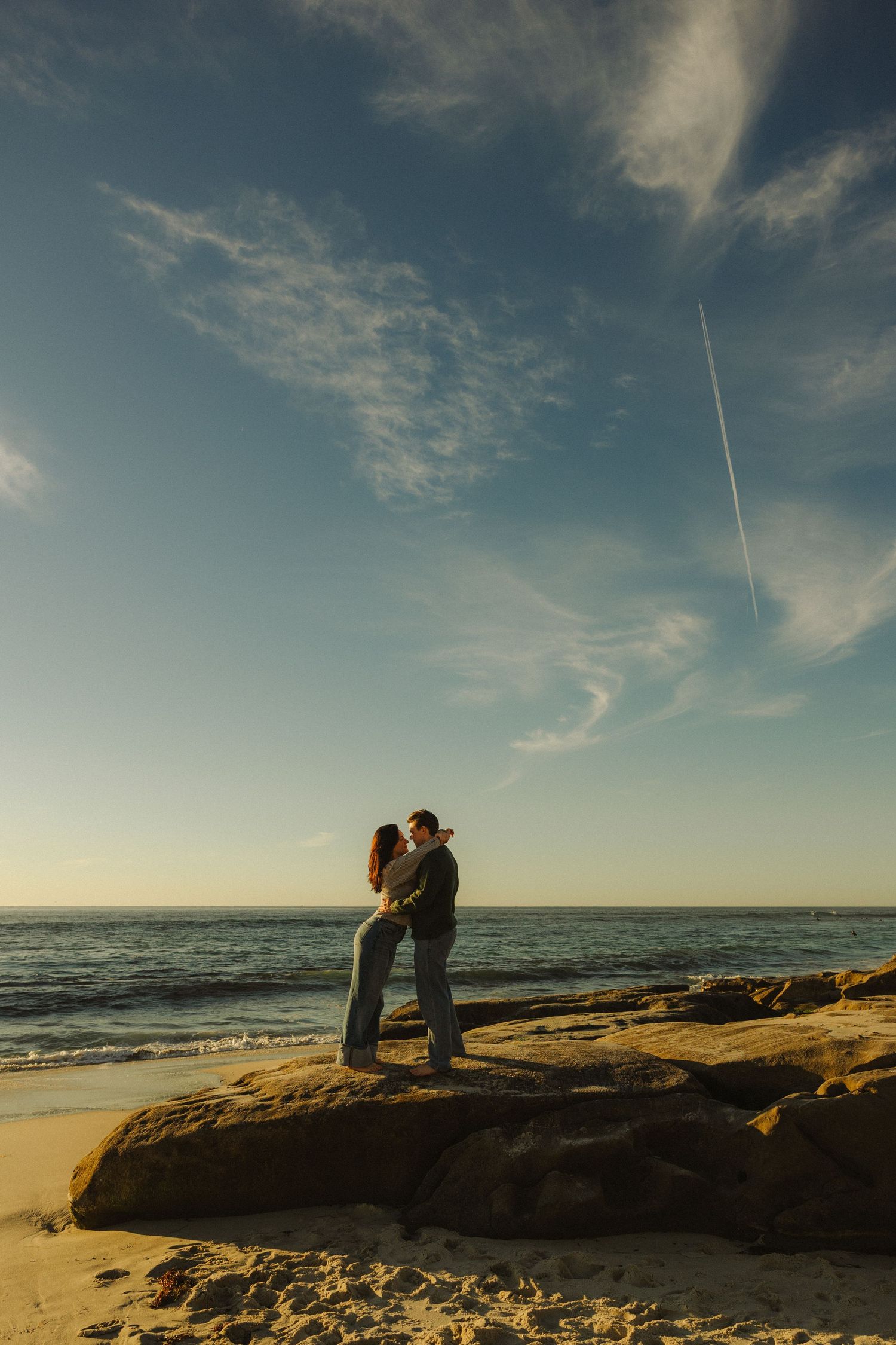 Two silhouettes embrace on a rocky beach cliff at sunset with ocean waves and dramatic clouds in the background.