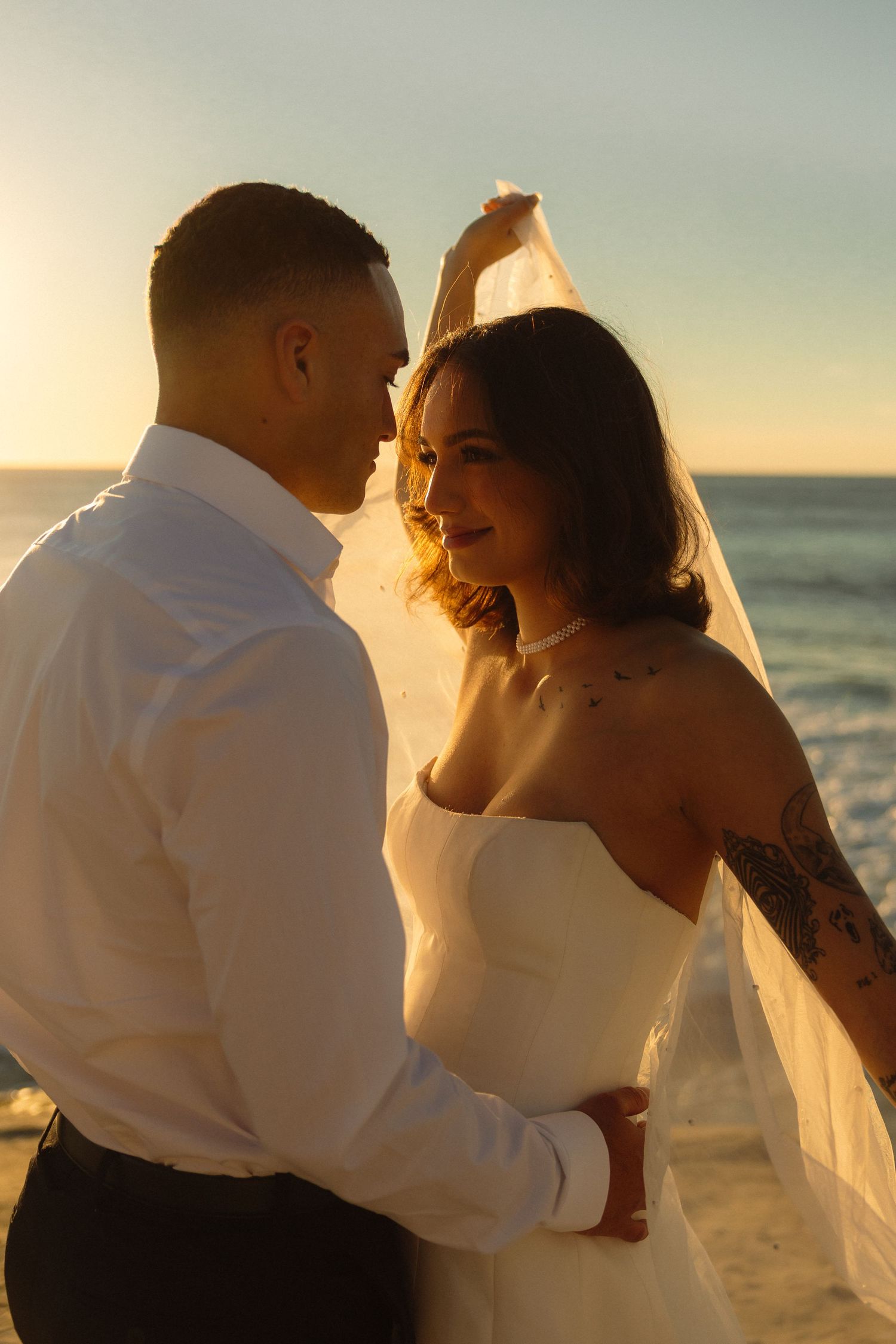 Newlywed couple shares intimate moment at sunset on beach with bride in white dress and groom in white shirt.