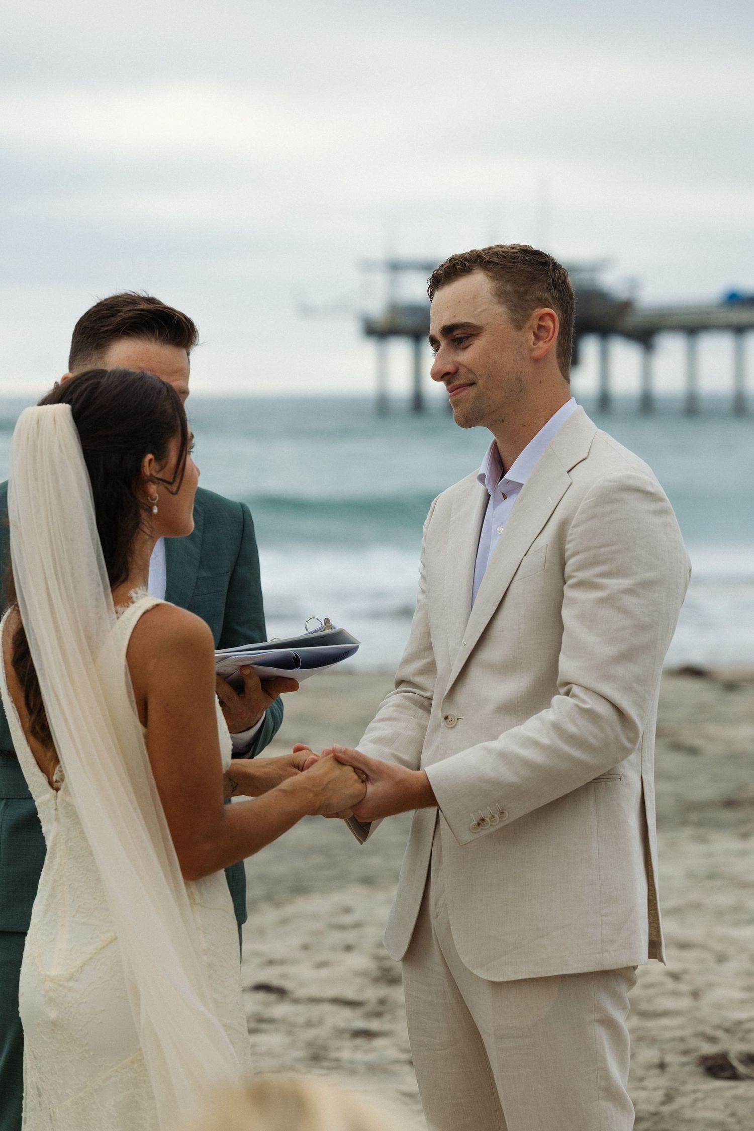 Couple exchanging vows at a beach wedding ceremony with an officiant and ocean pier backdrop.