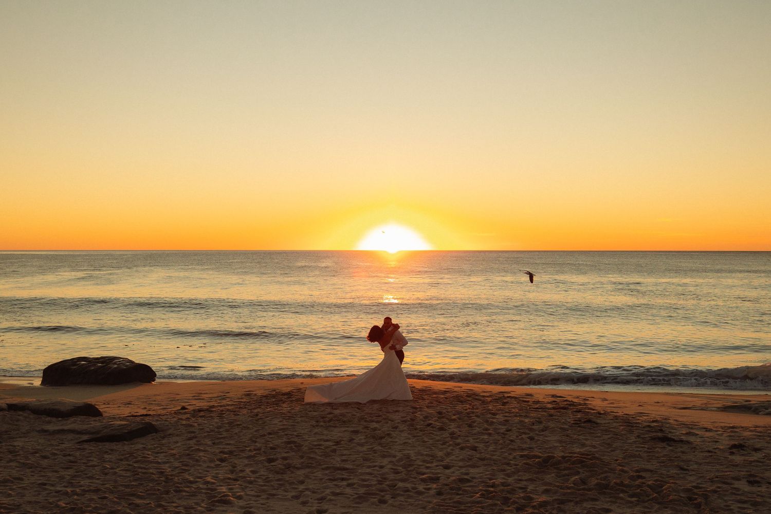 A silhouetted figure sits on a beach at sunset while waves roll in and the sun dips into the orange horizon.