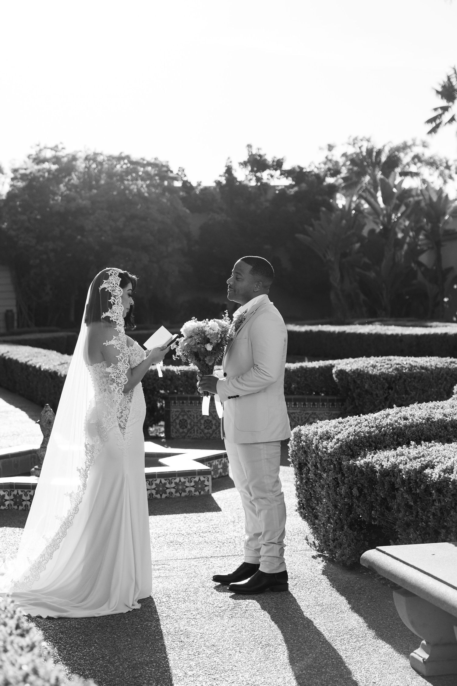 Black and white photo of a bride reading vows to groom in a lush garden setting during an outdoor wedding ceremony.