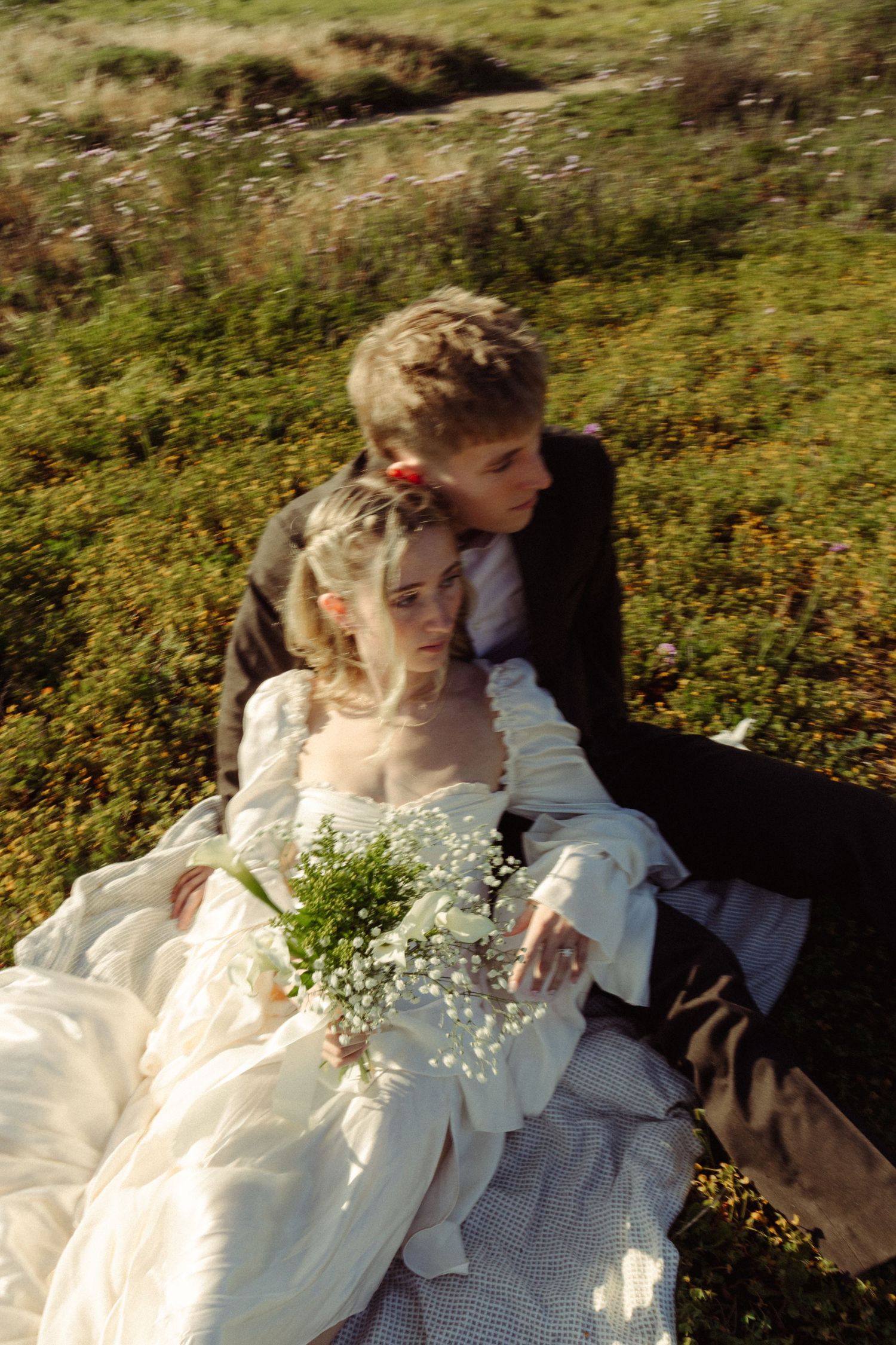 A bride in a white gown holds baby's breath flowers while sitting with a groom in a green meadow.