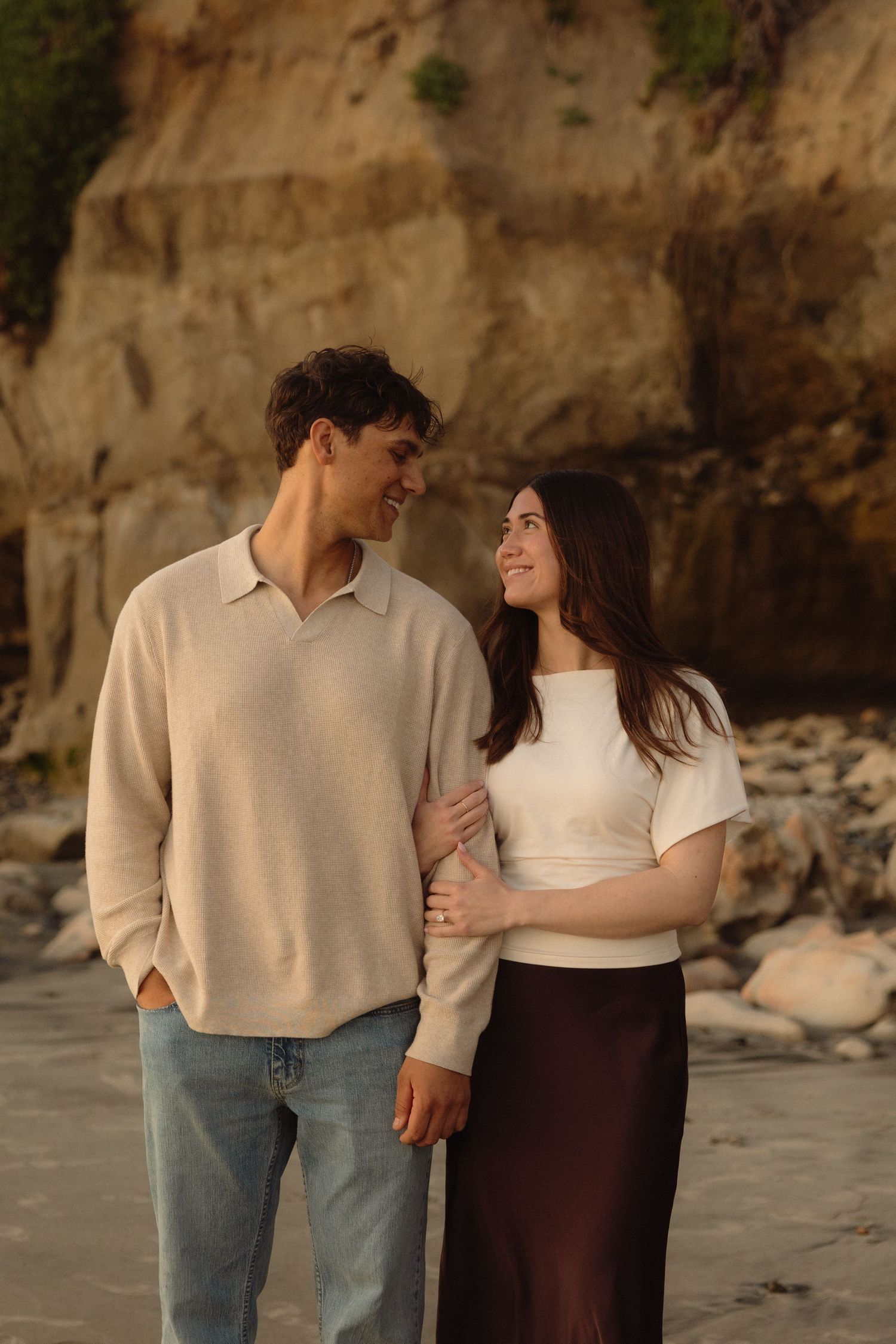 A couple in casual attire shares a tender moment against a backdrop of rocky cliffs at the beach.