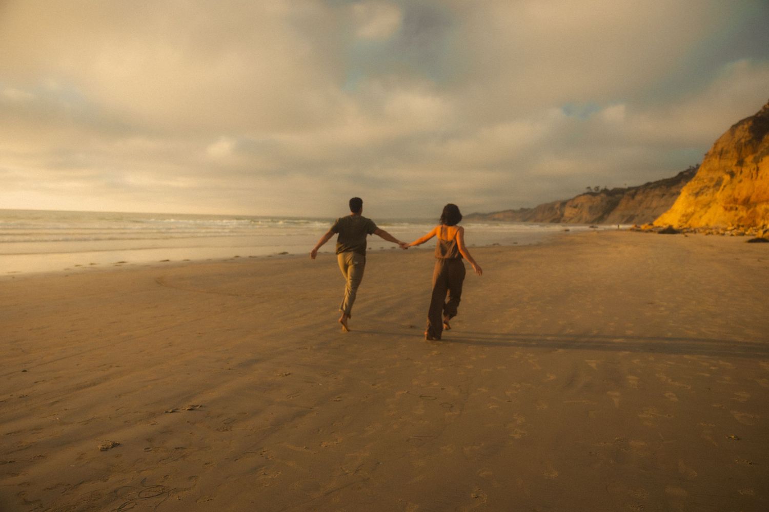 People and dogs play and run along a misty beach at sunset with dramatic cliffs in the background.