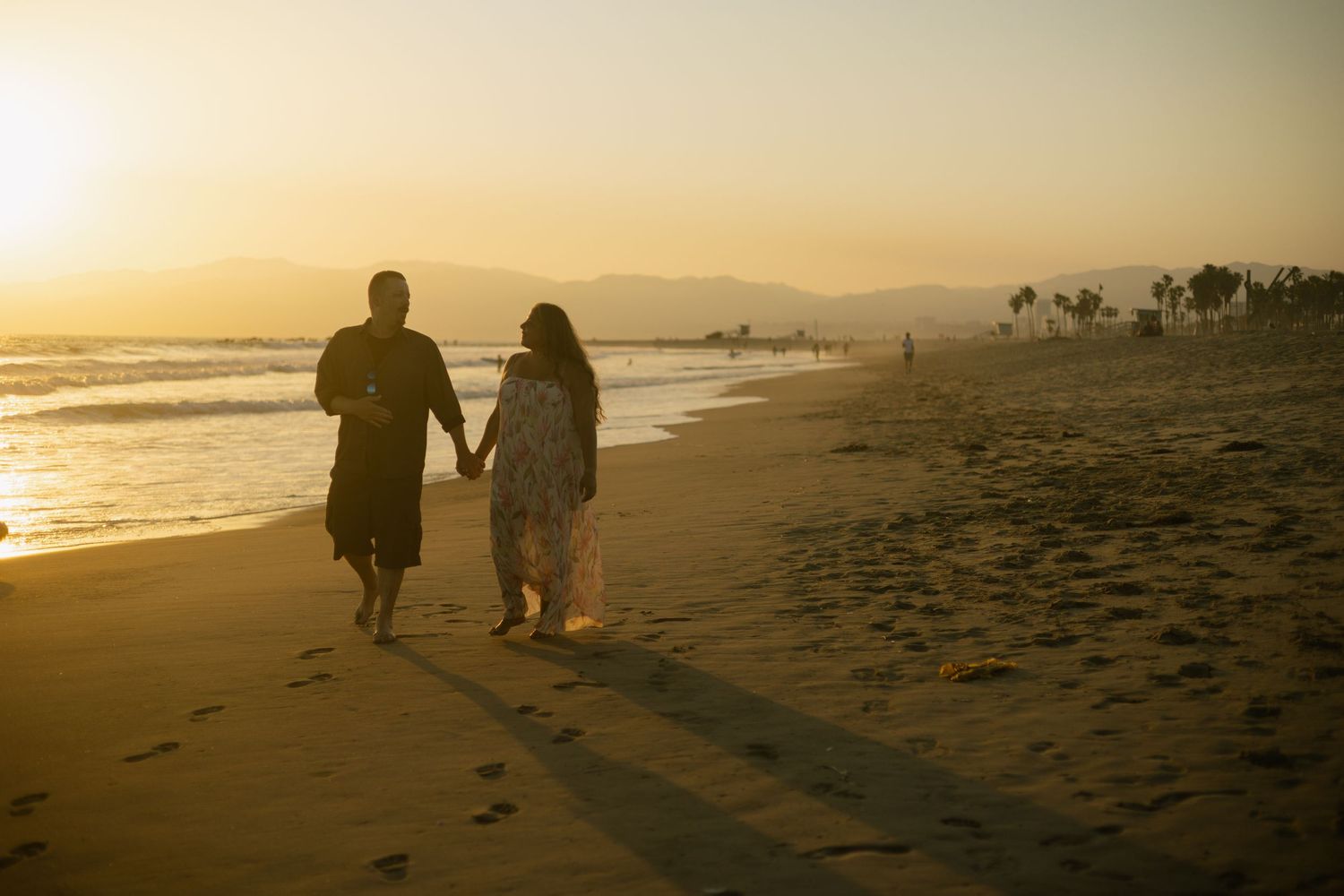 Couple walking together on sandy beach at sunset with palm trees and waves in the golden light.