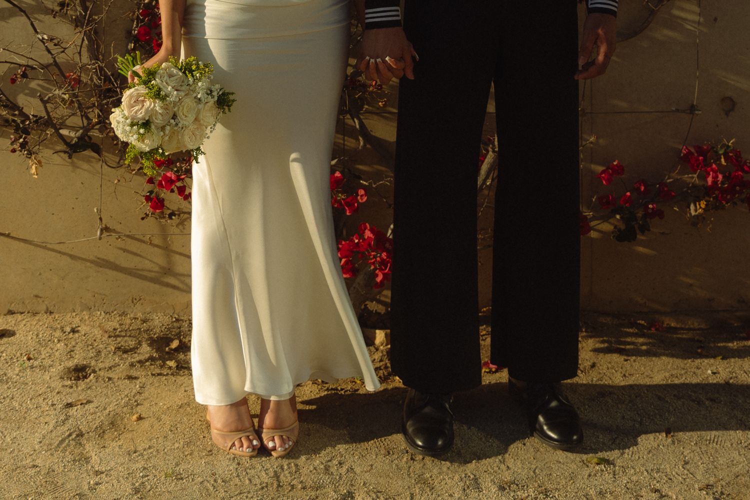 Two pairs of feet stand close together against a wall with red flowers, one wearing a white dress and tan shoes.