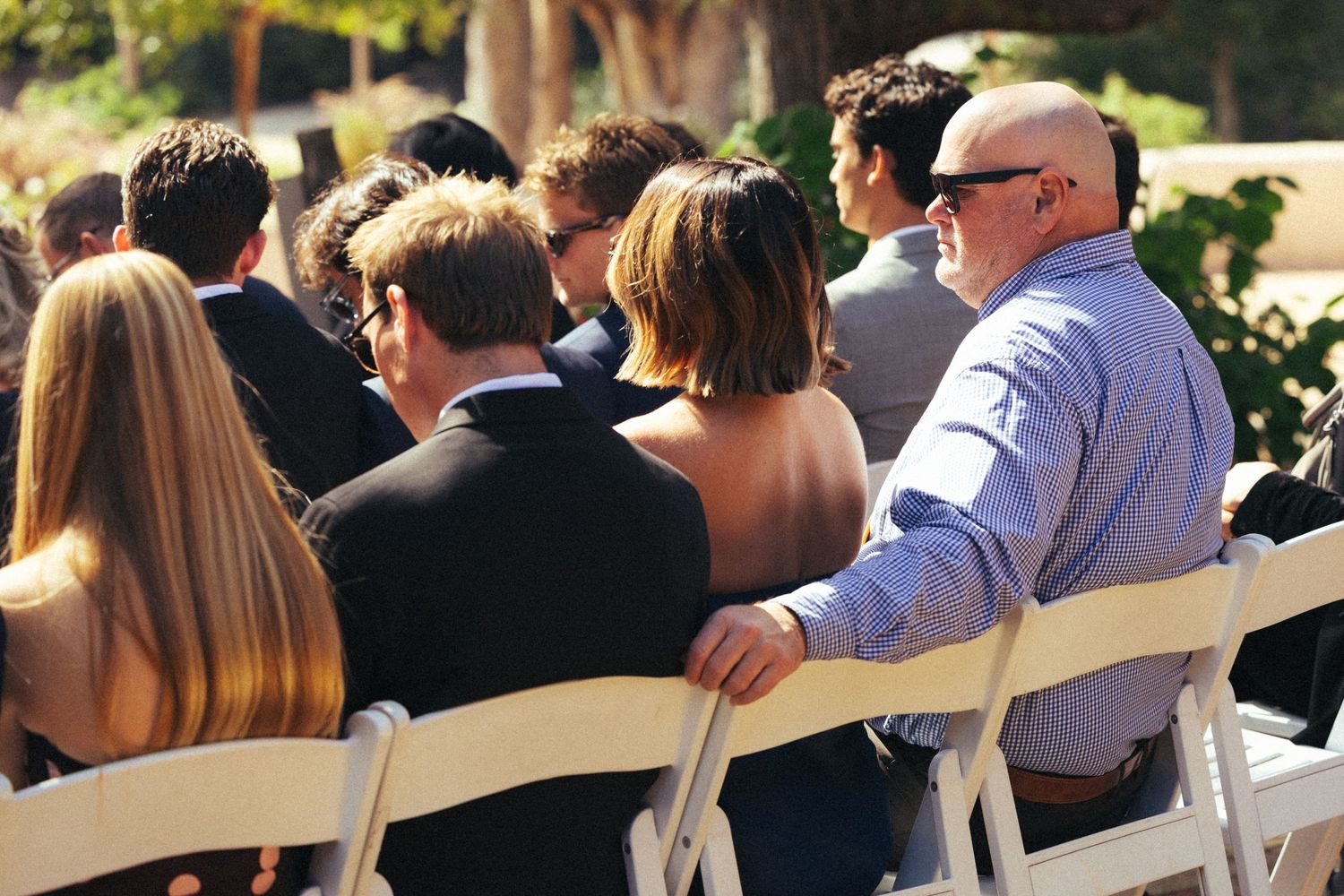 Guests seated in white chairs at an outdoor wedding ceremony, dressed in formal attire on a sunny day.