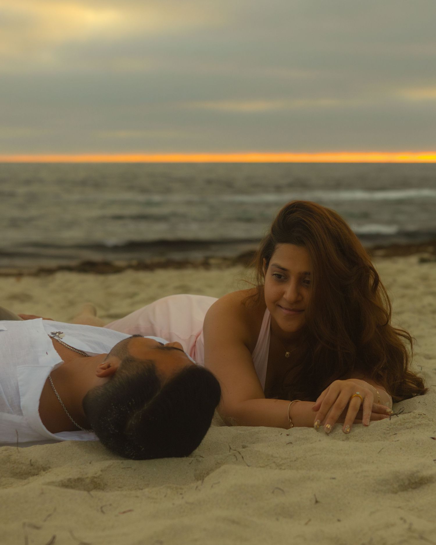 A couple relaxes together on a sandy beach at sunset with the ocean and golden sky in the background.