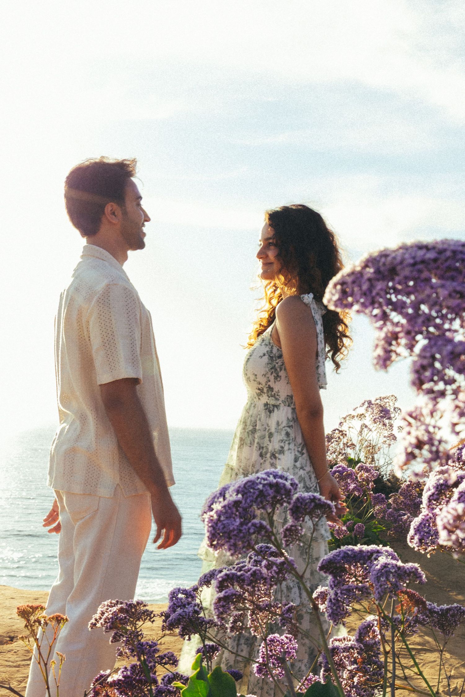 Couple standing among purple wildflowers by the ocean, smiling at each other in warm sunlight.