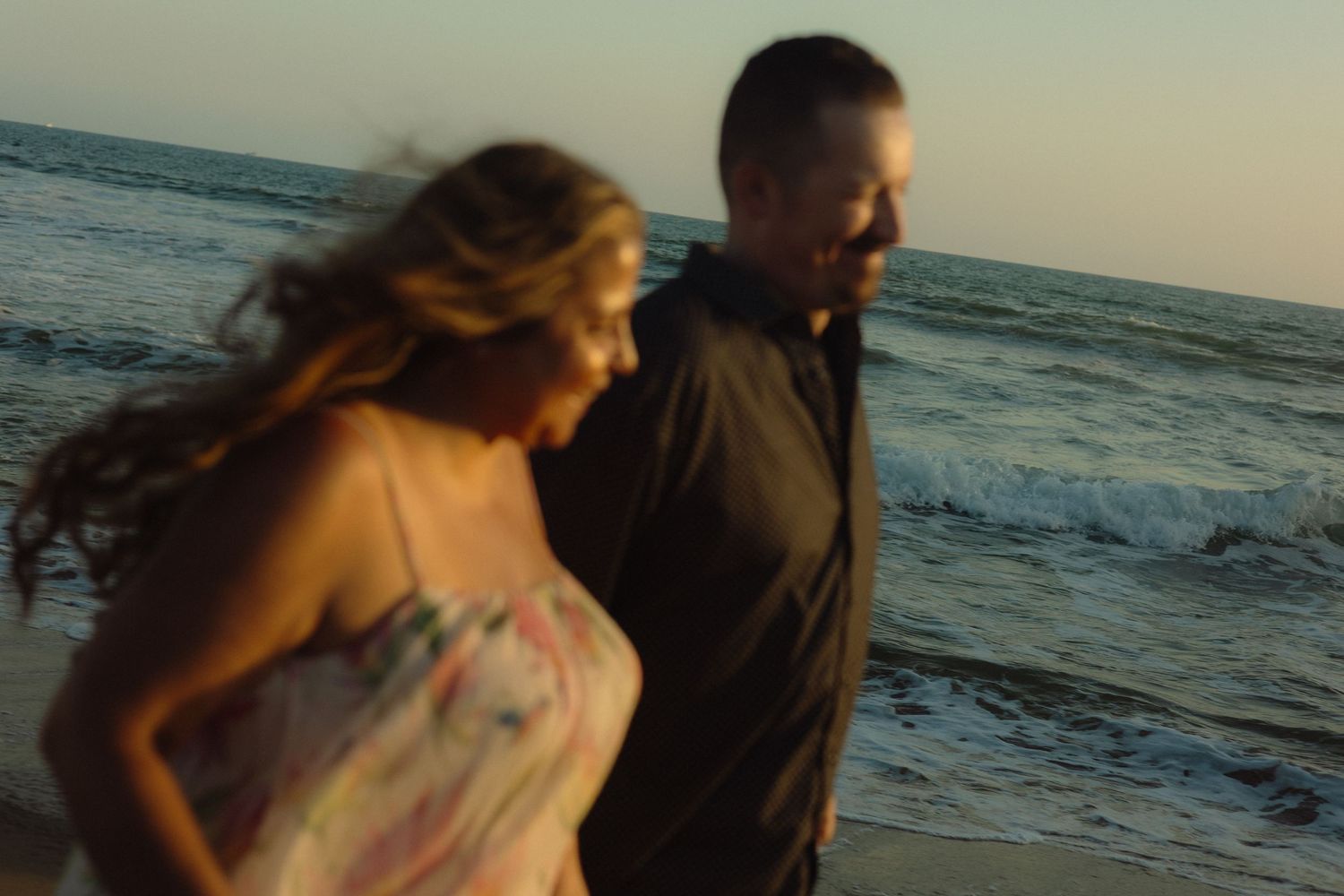 A couple takes a romantic stroll along the beach at sunset with waves crashing behind them.